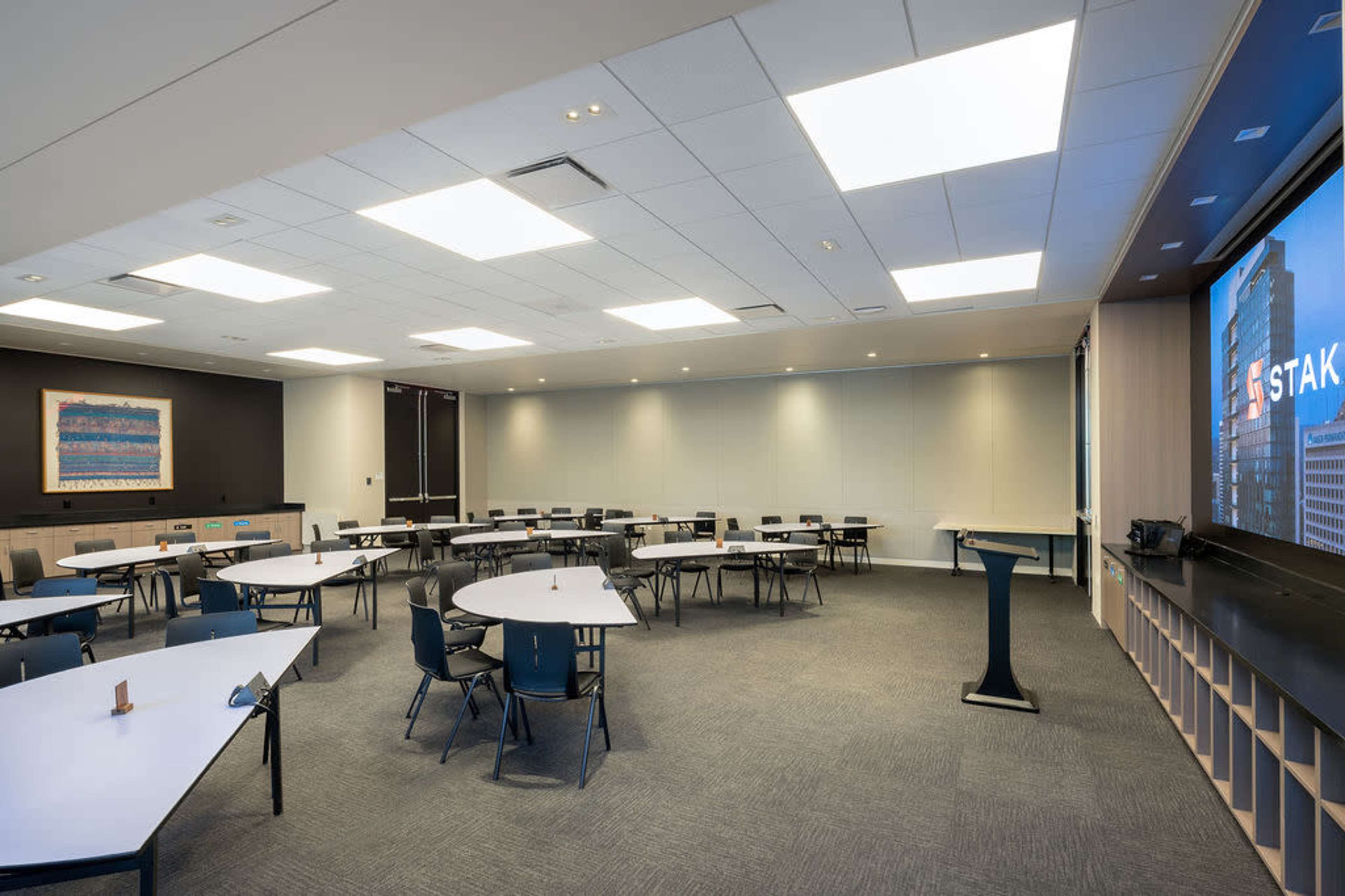The image shows a spacious conference room set up with several round tables and chairs, along with a presentation screen and podium.