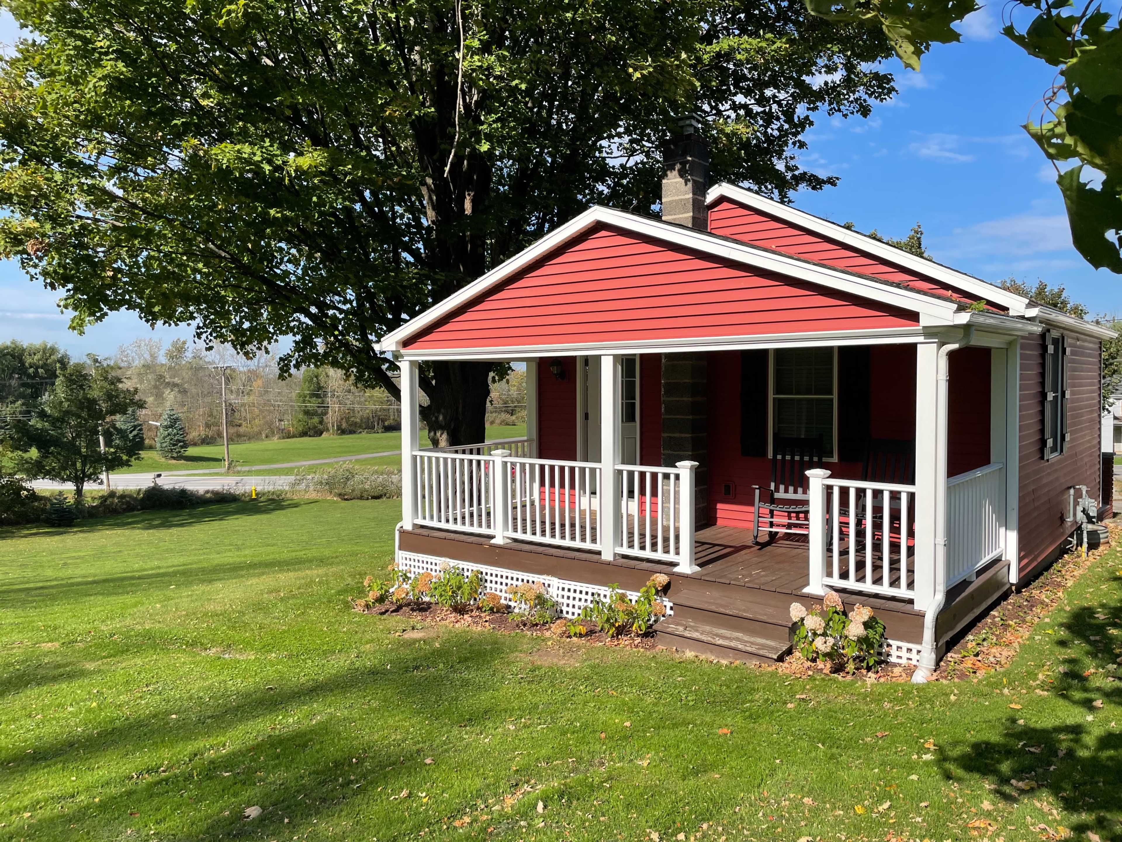 A small red house with a white porch and railing sits on a grassy lawn surrounded by trees.