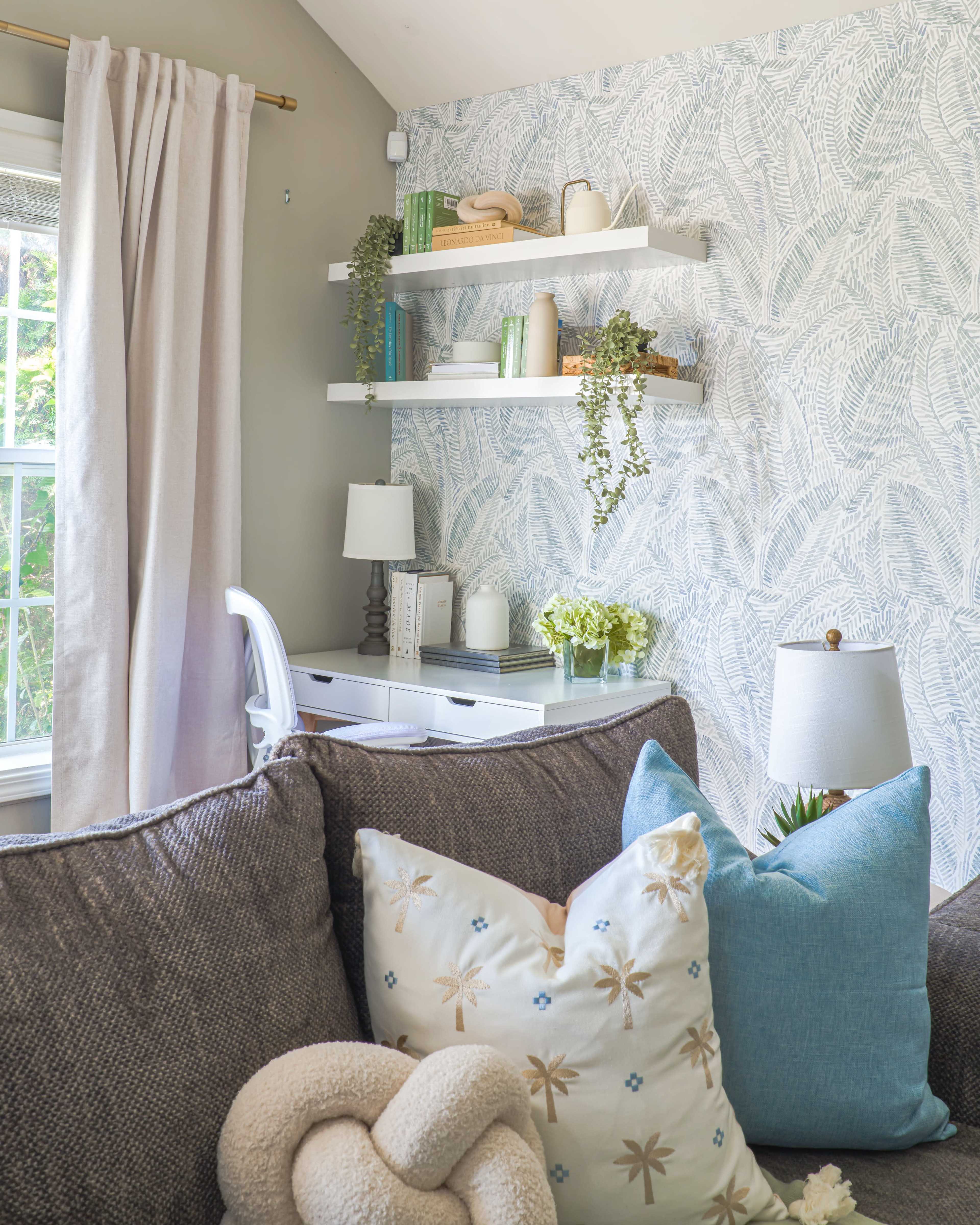 A cozy room features a sectional sofa adorned with decorative pillows, a desk with shelves above displaying plants and decor, and a wall with patterned wallpaper.
