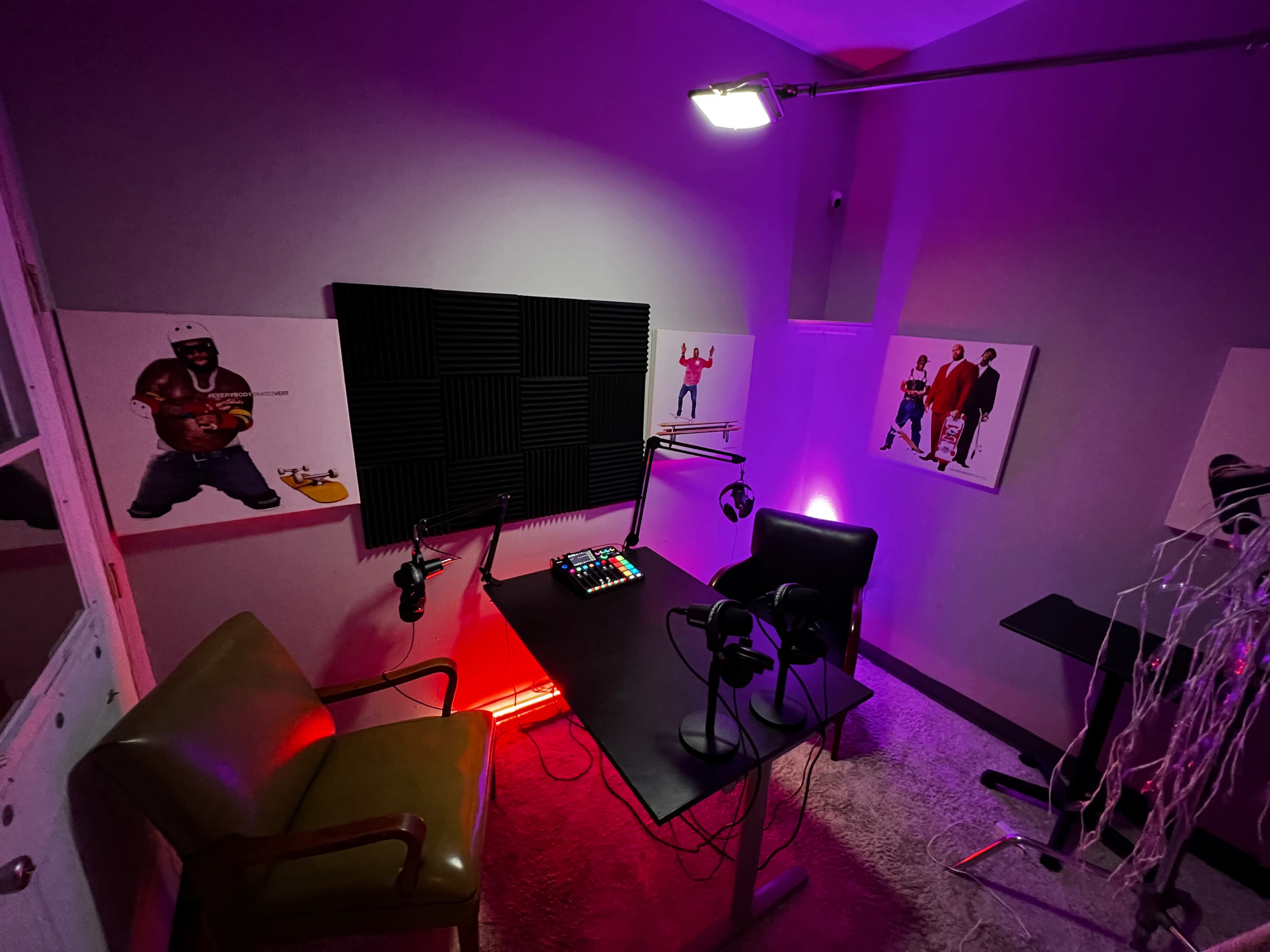 A recording studio features a table with microphones, headphones, and colorful lighting, surrounded by soundproof panels and artwork on the walls.