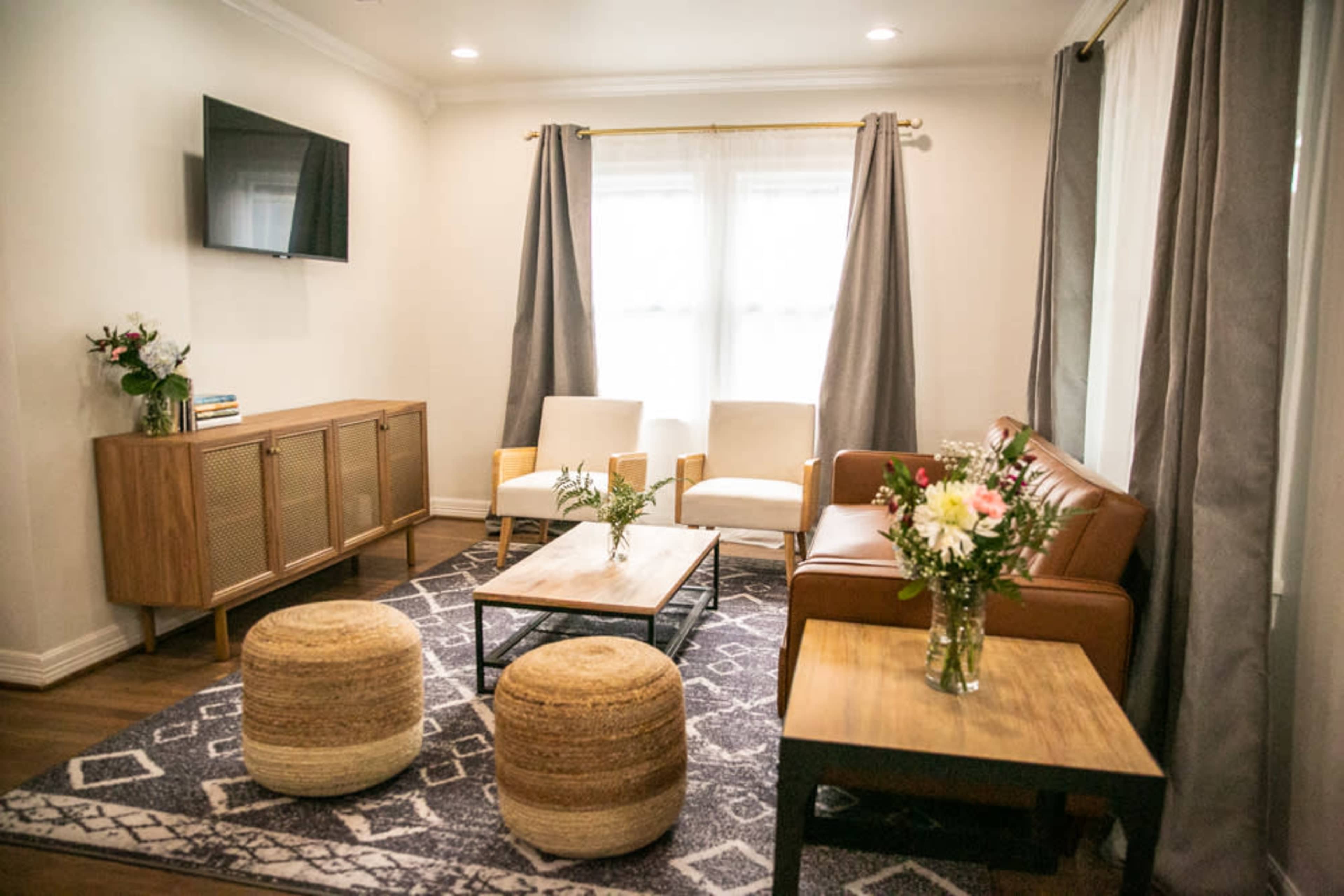 The living room features a television mounted on the wall, two white chairs, a brown leather sofa, a coffee table, and a patterned rug, surrounded by large windows with curtains.