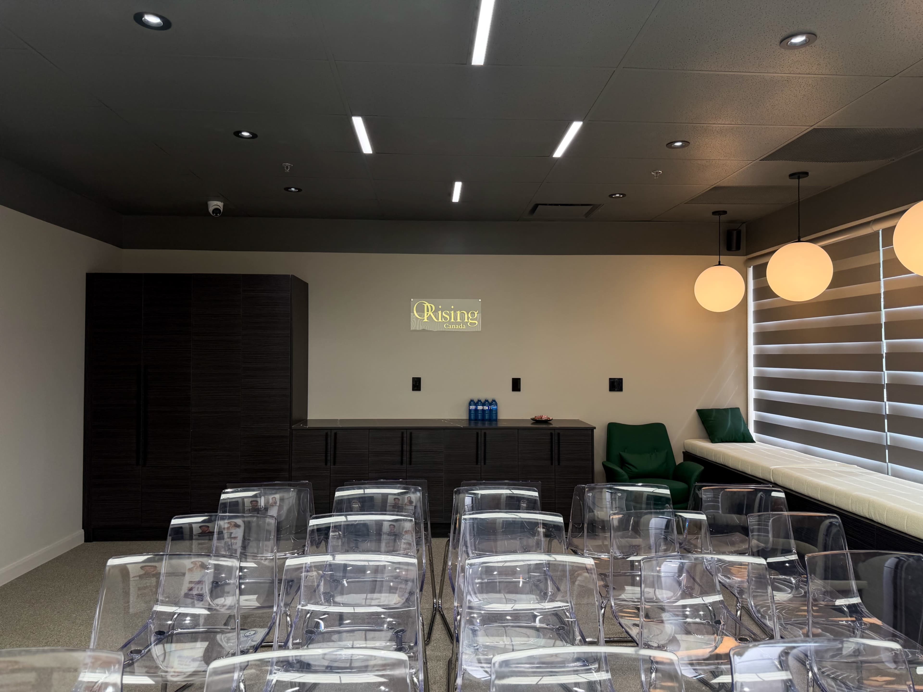A modern meeting room features transparent chairs arranged in rows facing a dark cabinet, with a wall displaying the word "Rising" illuminated above it and two large round light fixtures hanging from the ceiling.