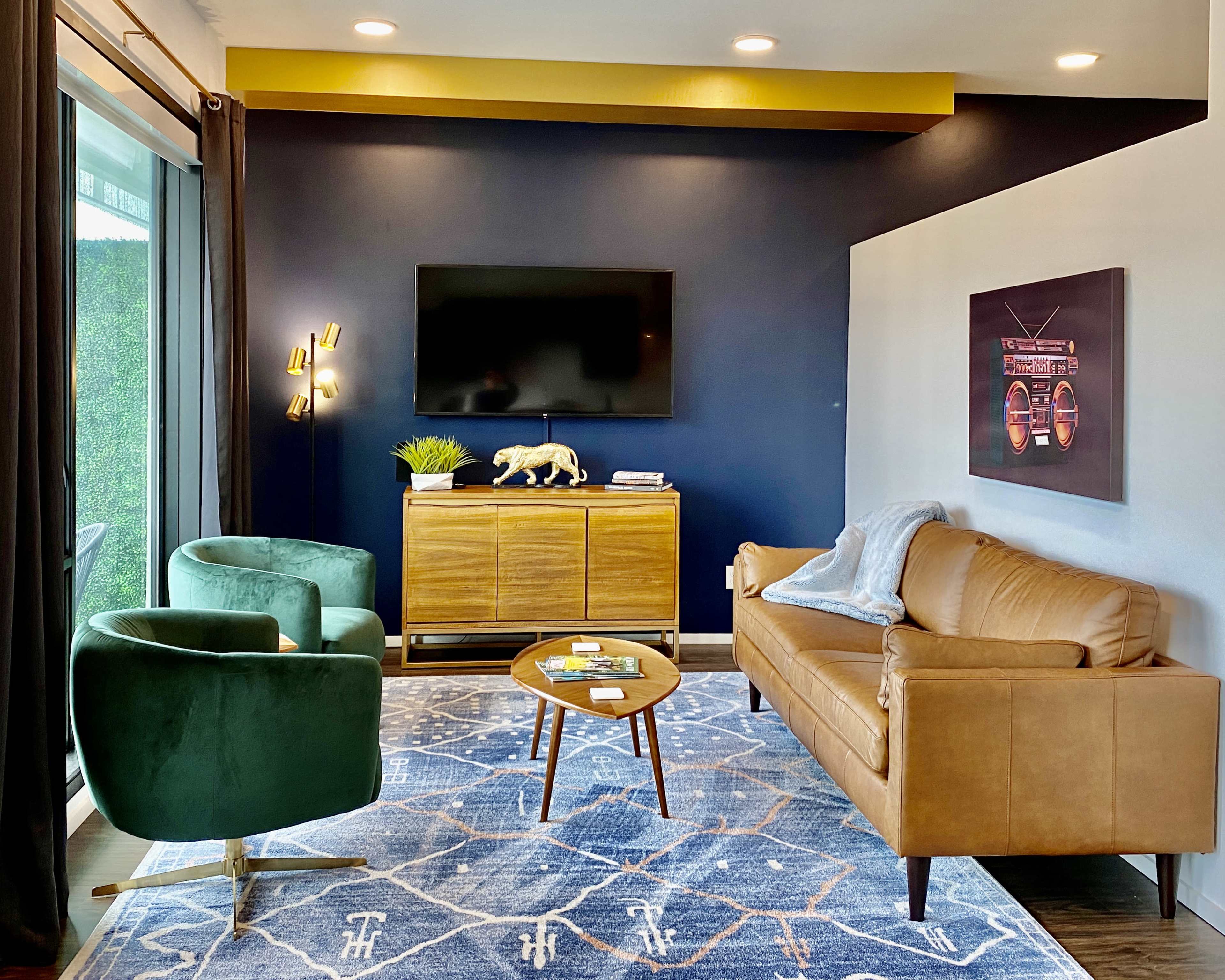 The living room features a leather sofa, two green armchairs, a wooden sideboard, a television mounted on the wall, and a patterned area rug.