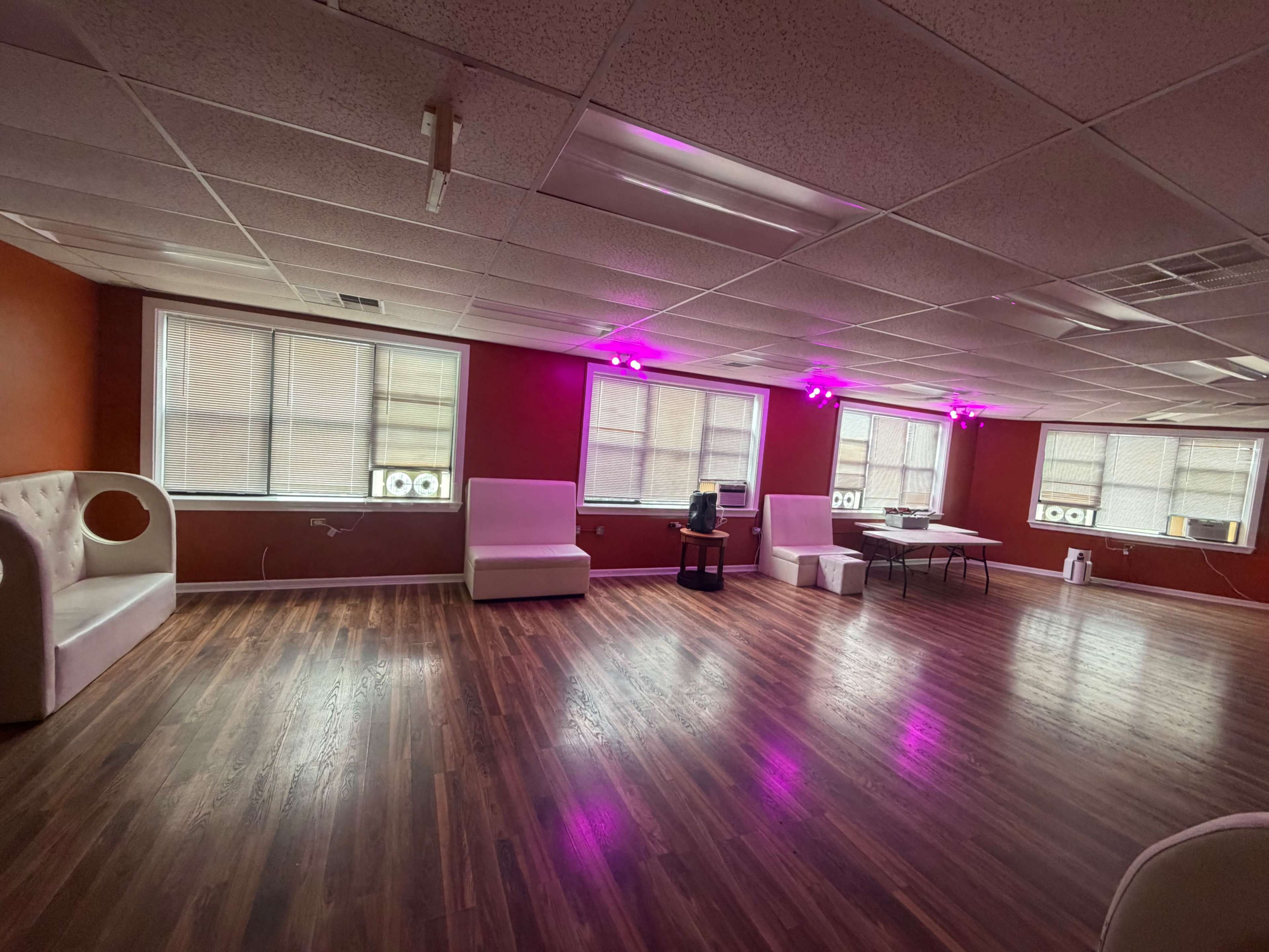 The image shows a spacious room with wooden flooring, white couches, a table in the center, and pink lights on the ceiling.