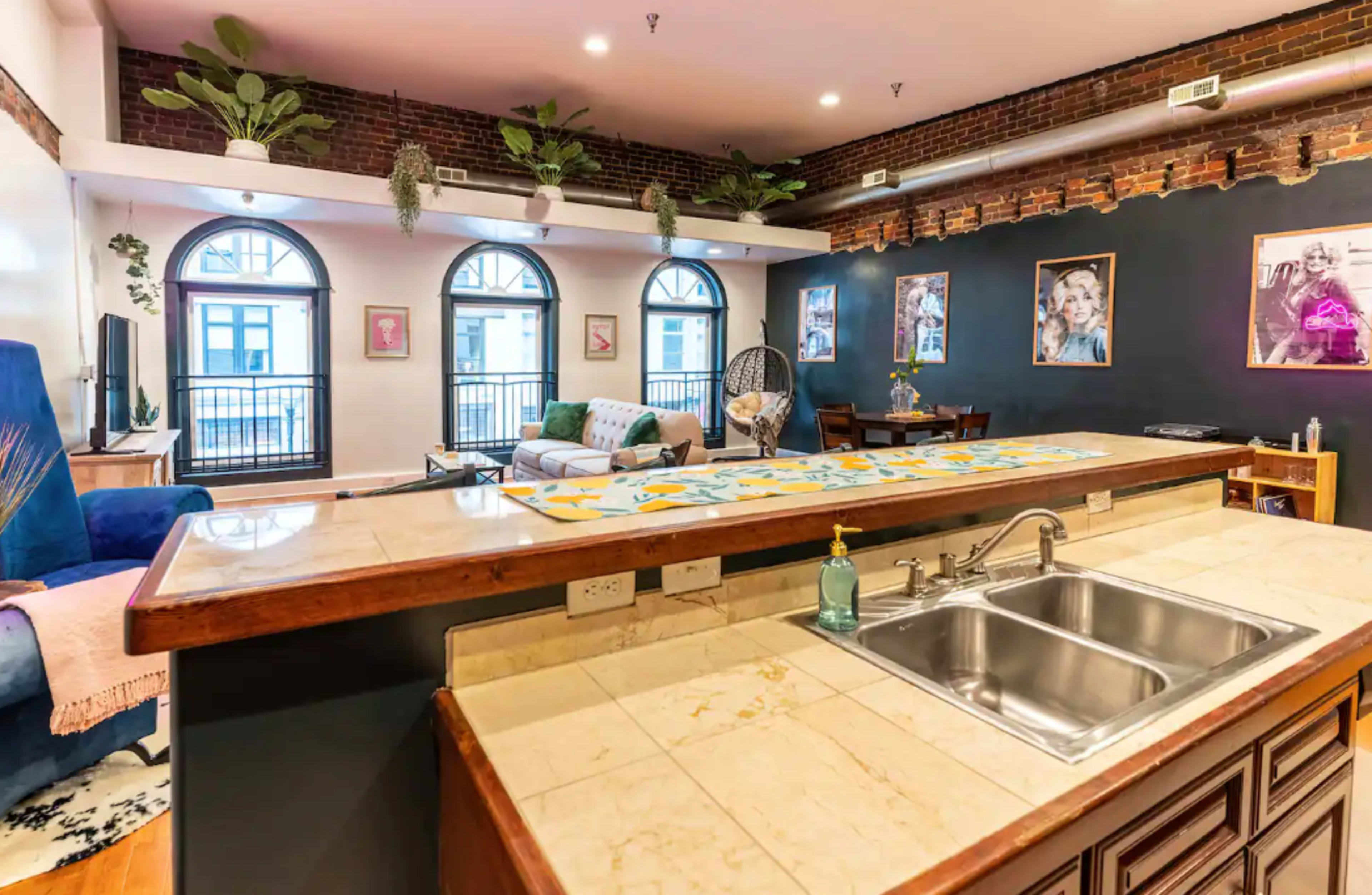 A spacious living area with exposed brick walls, large windows, and a kitchen island with a sink in the foreground.
