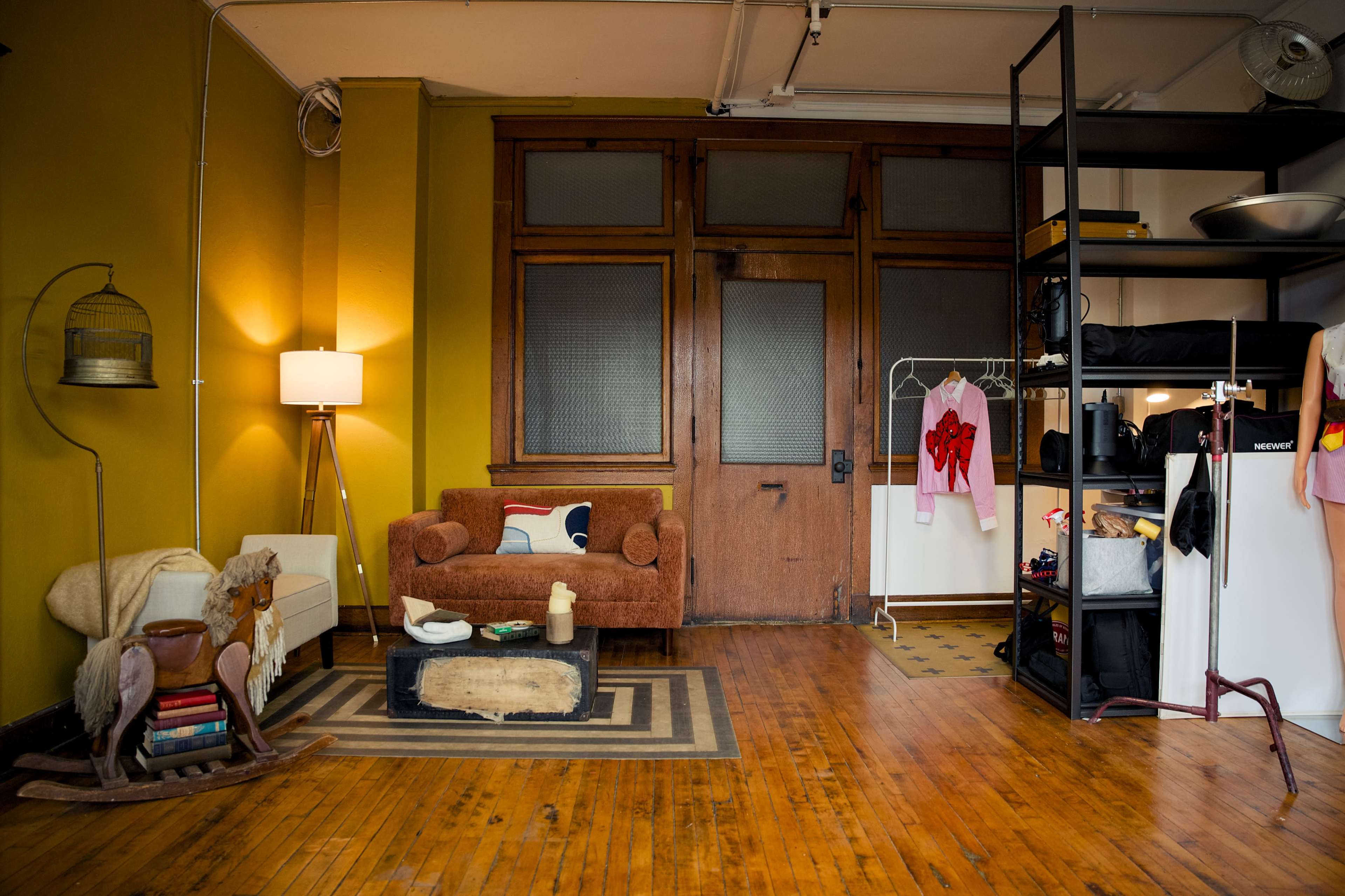 The image shows a cozy living space with a brown couch, books on a small table, and a shelving unit displaying various items, alongside a large wooden door and a warm yellow wall.
