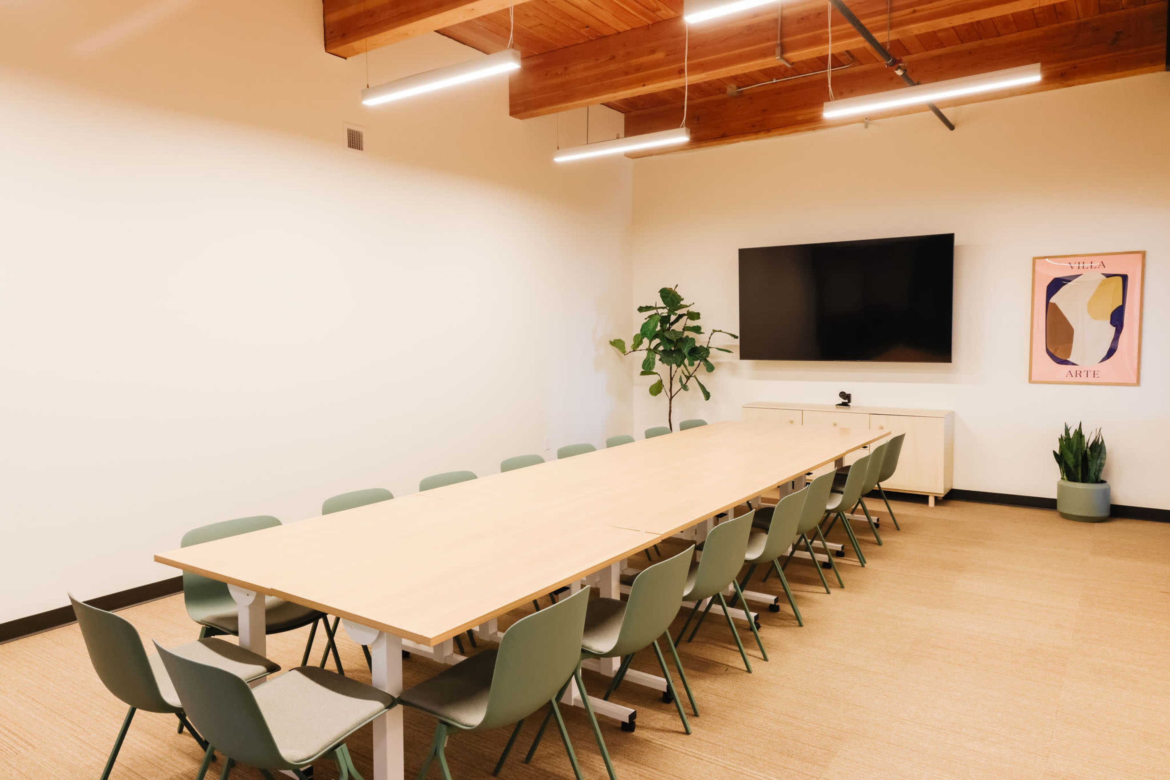 A modern conference room features a long wooden table surrounded by green chairs, with a television mounted on the wall and a plant in the corner.