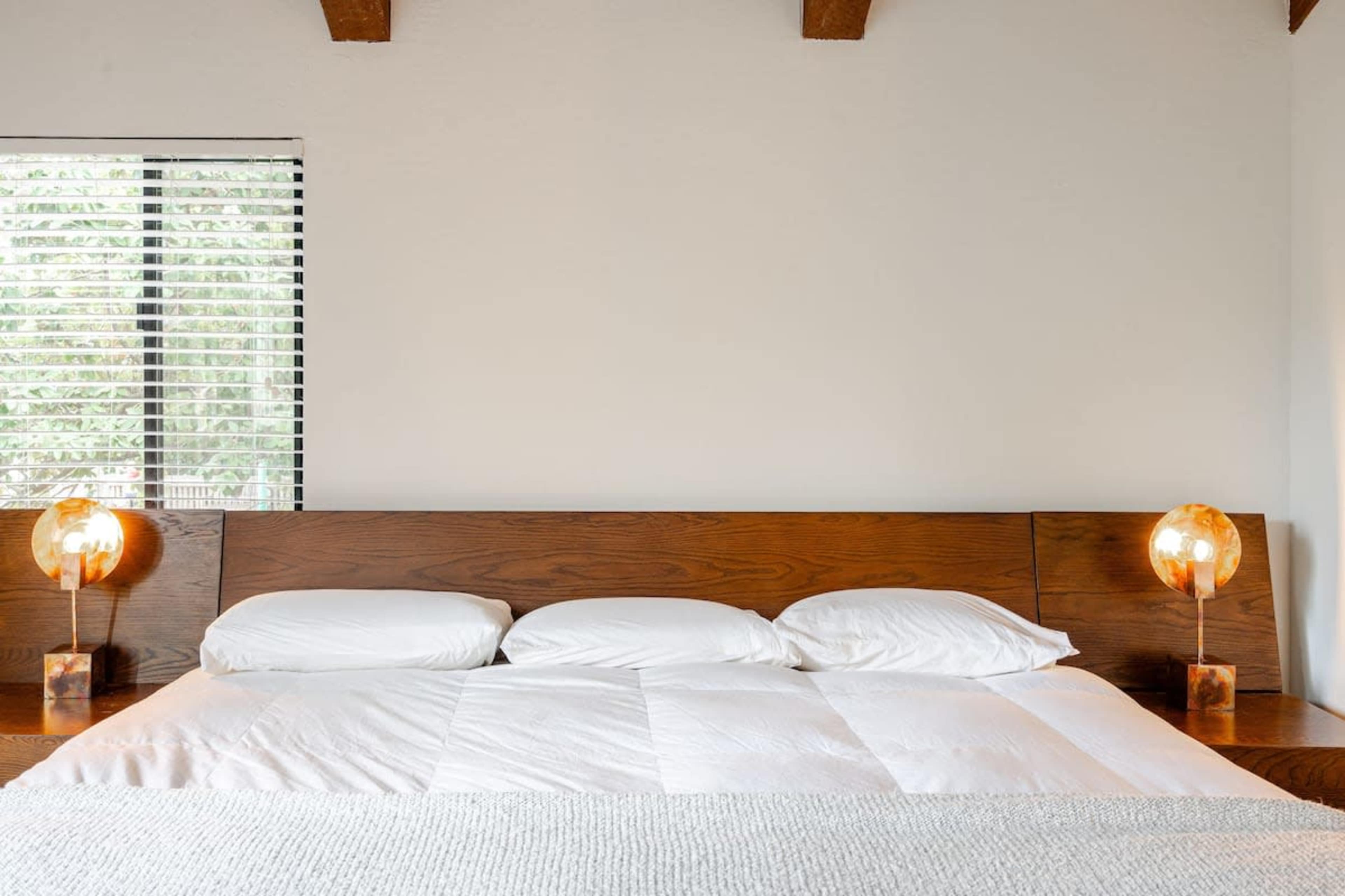 A simple bedroom scene features a wooden bed with three white pillows and two illuminated lamps on either side, against a wall with a window covered by blinds.