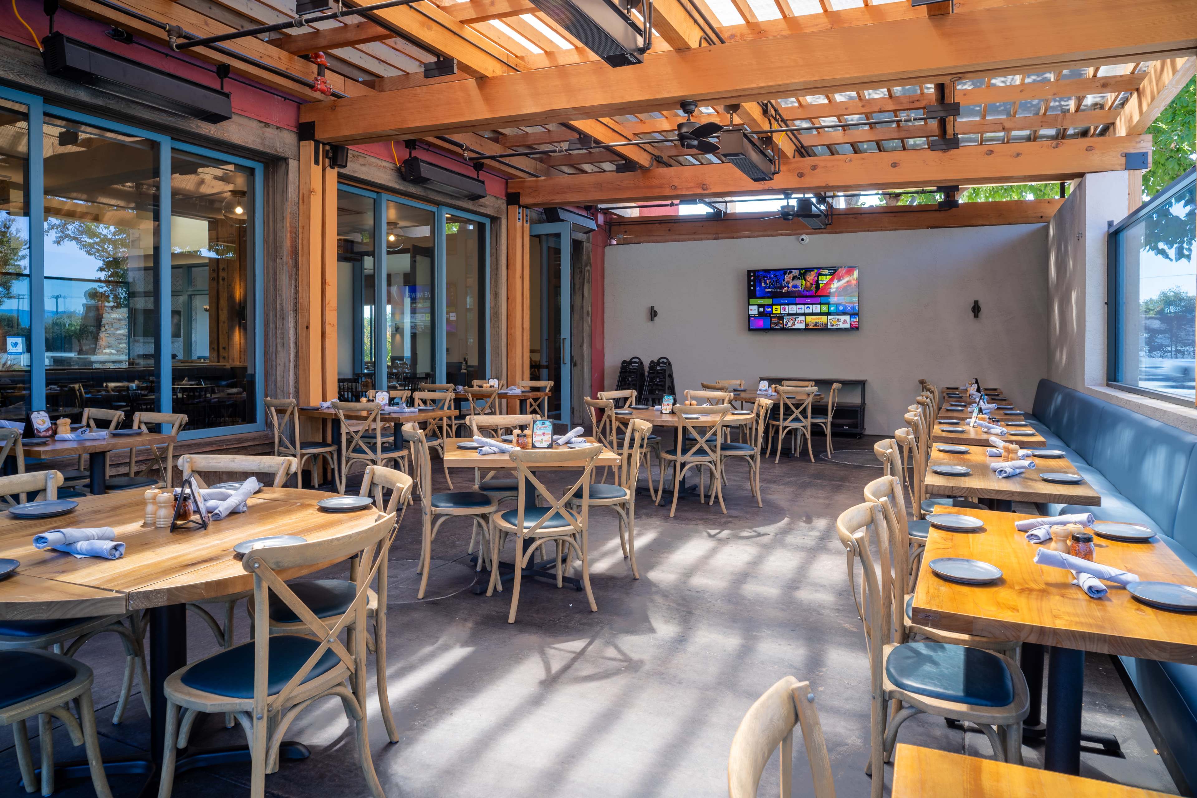 The image shows a spacious restaurant interior with wooden tables and chairs, featuring large windows and a television mounted on the wall.