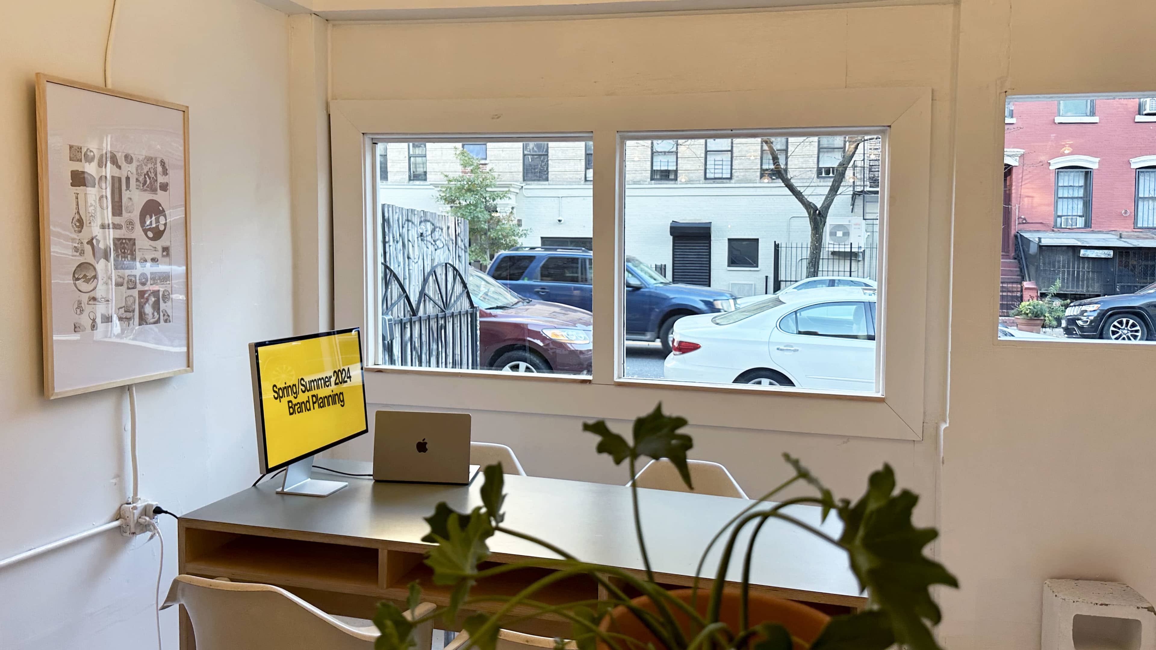 A bright workspace with a table, a computer monitor displaying "Spring/Summer 2024 Brand Drawing," and large windows overlooking a street with parked cars.