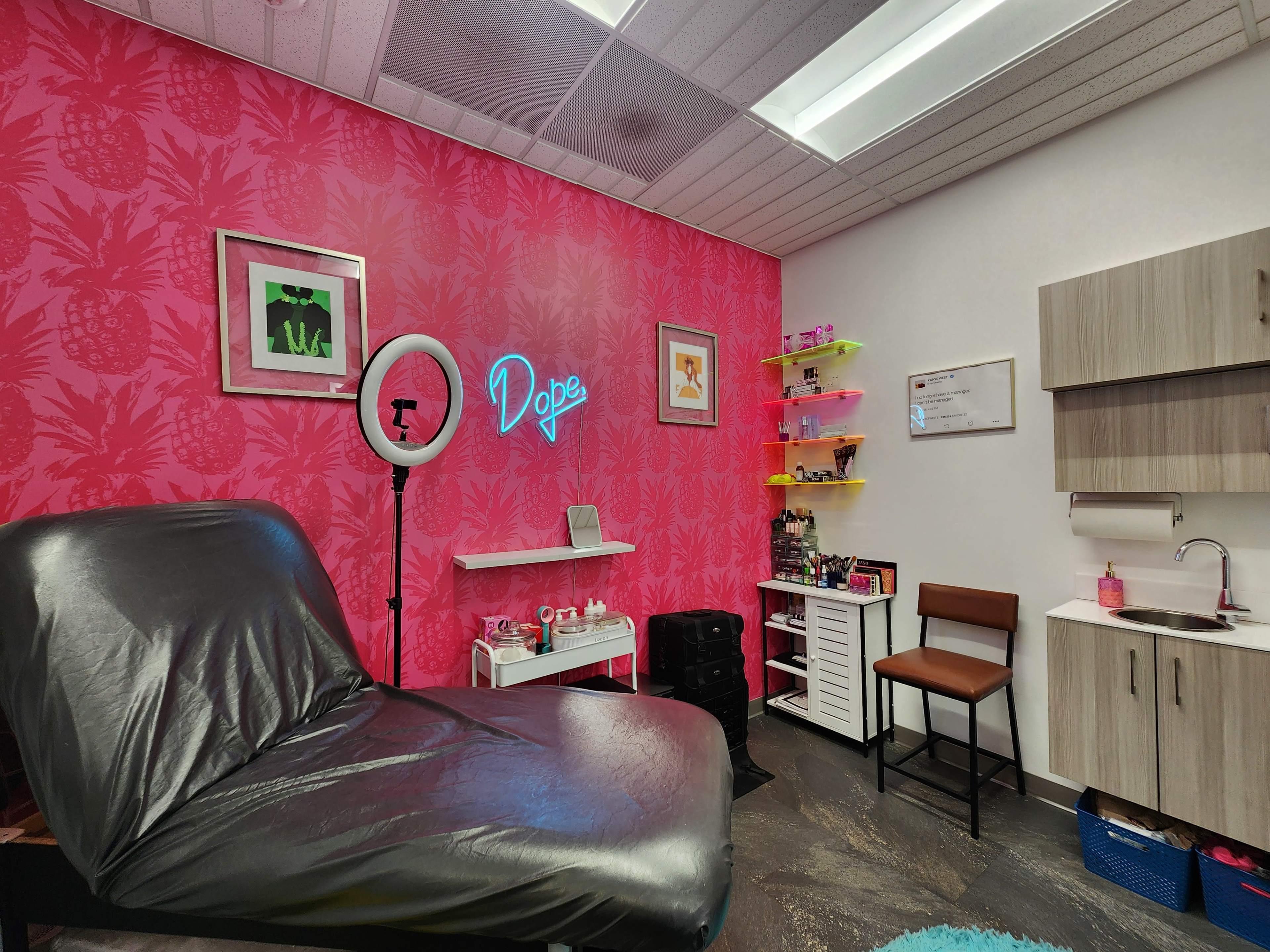 The image shows a vibrantly decorated tattoo studio featuring a black treatment chair, a neon sign that reads "Dope," and shelves displaying various beauty products against a pink tropical-patterned wall.