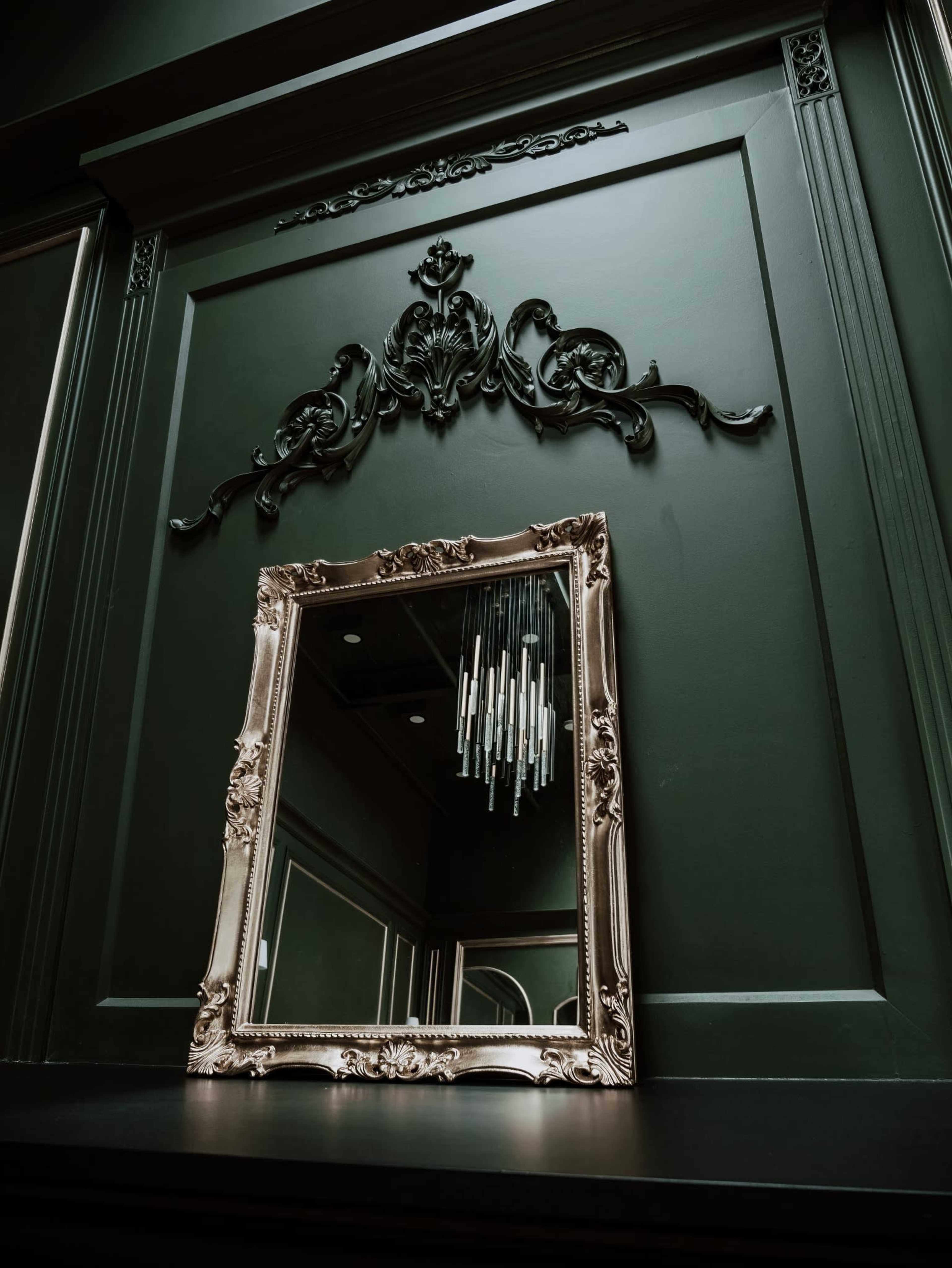A decorative mirror is framed against a dark green wall adorned with intricate molding and a modern light fixture above it.