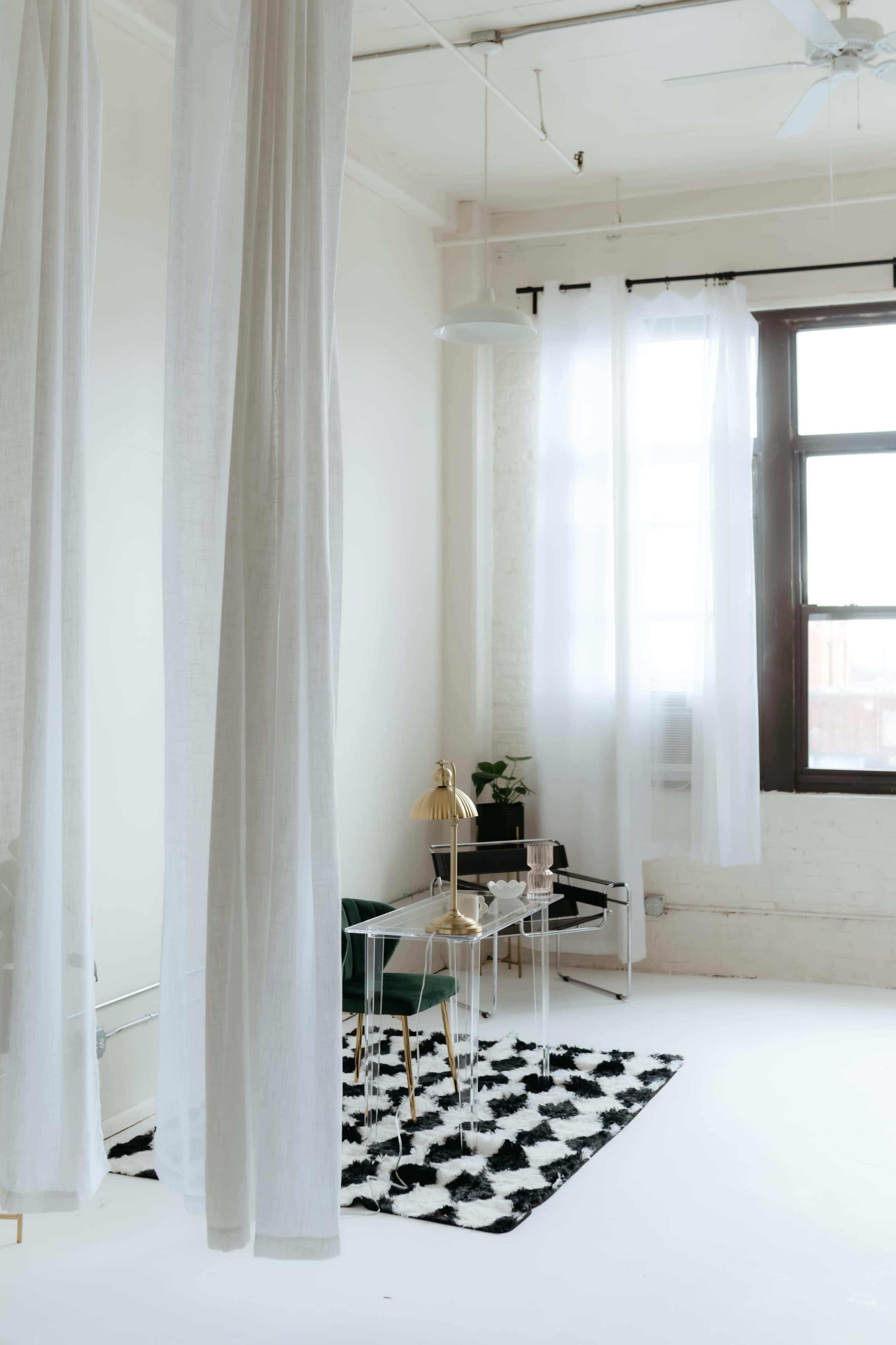 The image depicts a bright room with sheer white curtains, a small table with a lamp, and green chairs on a black and white rug.