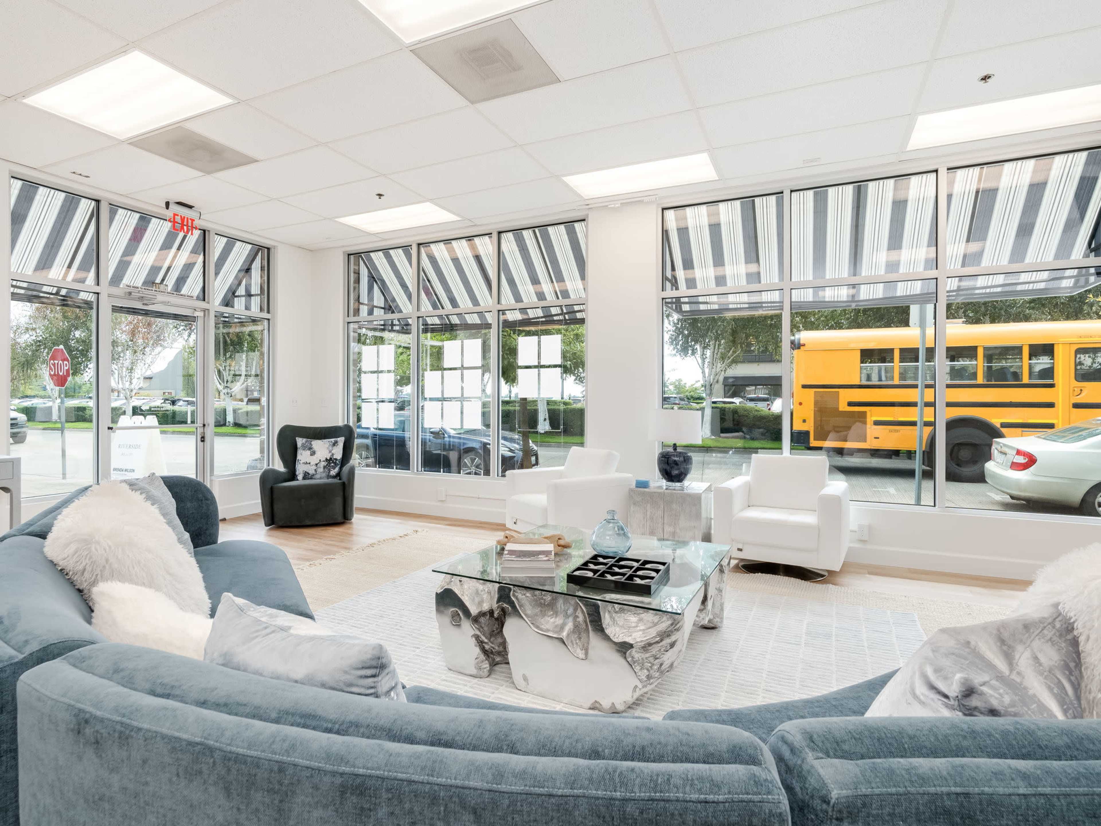 A brightly lit waiting area features a curved blue sofa, two white chairs, and large windows with a view of a yellow school bus parked outside.