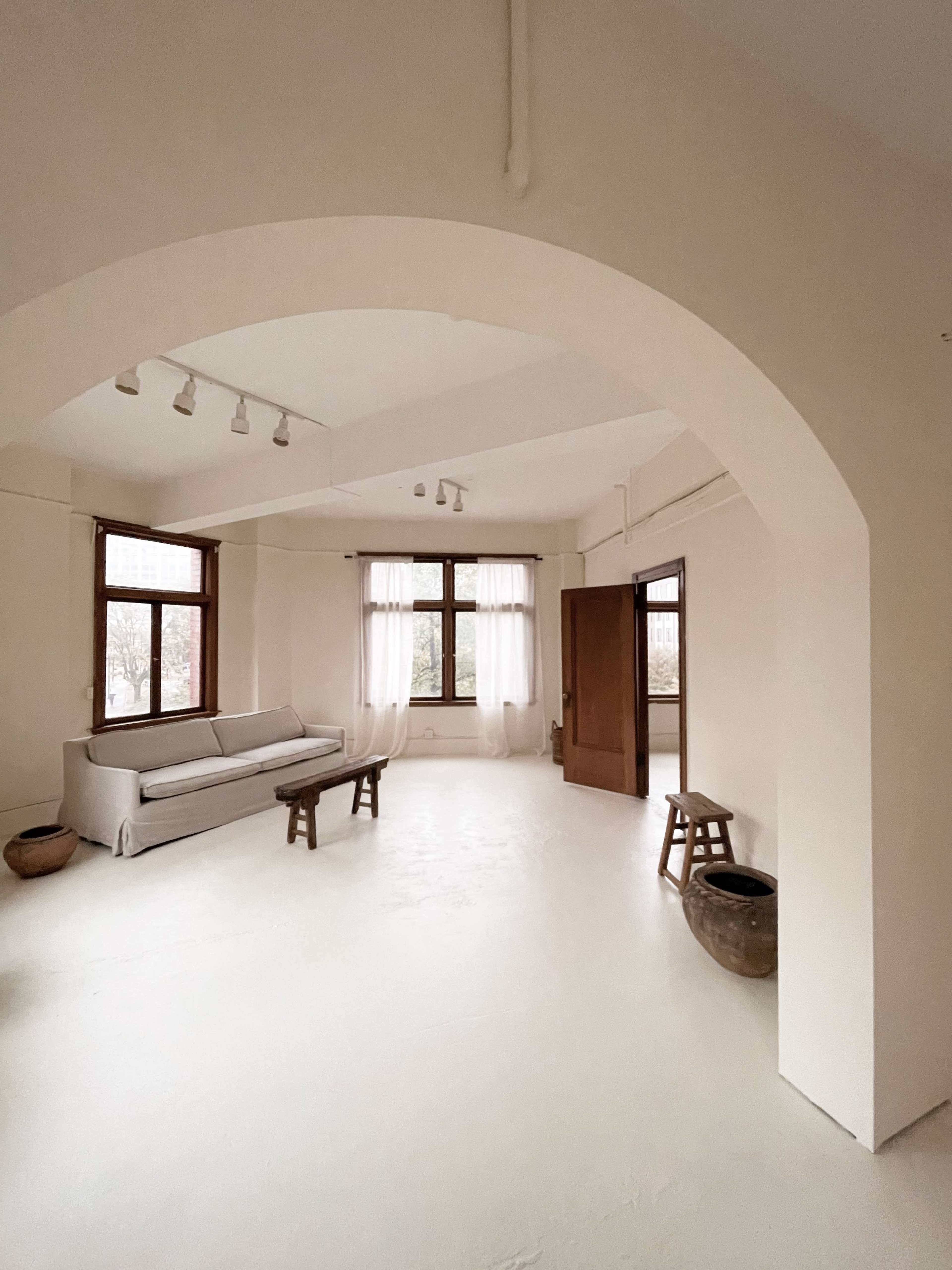An open, airy room features a light-colored couch, two chairs, large windows, and minimal furnishings.