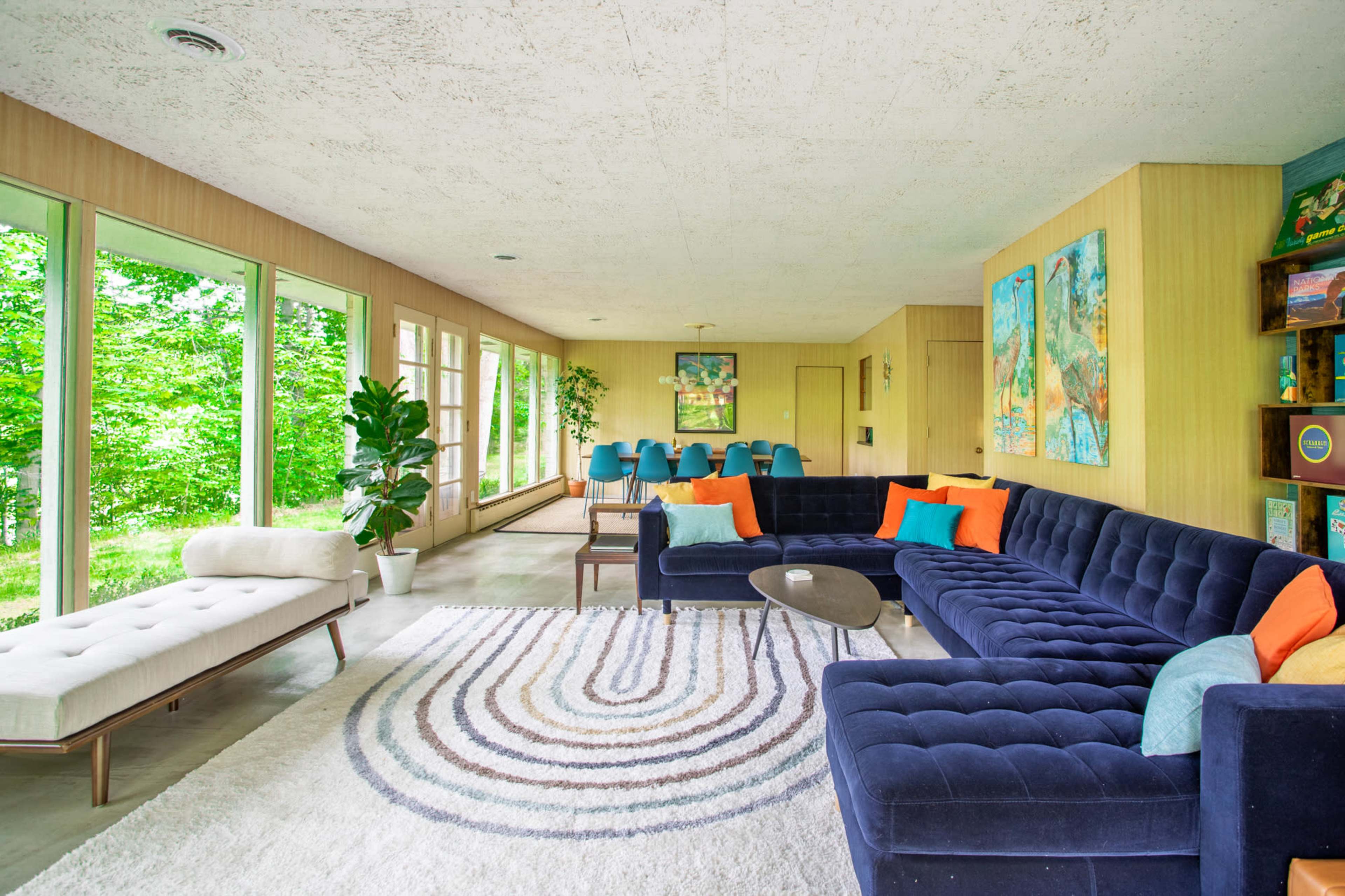 The room features a spacious living area with large windows, a blue sectional sofa, a round coffee table, and a dining area with blue chairs and a light wood table.