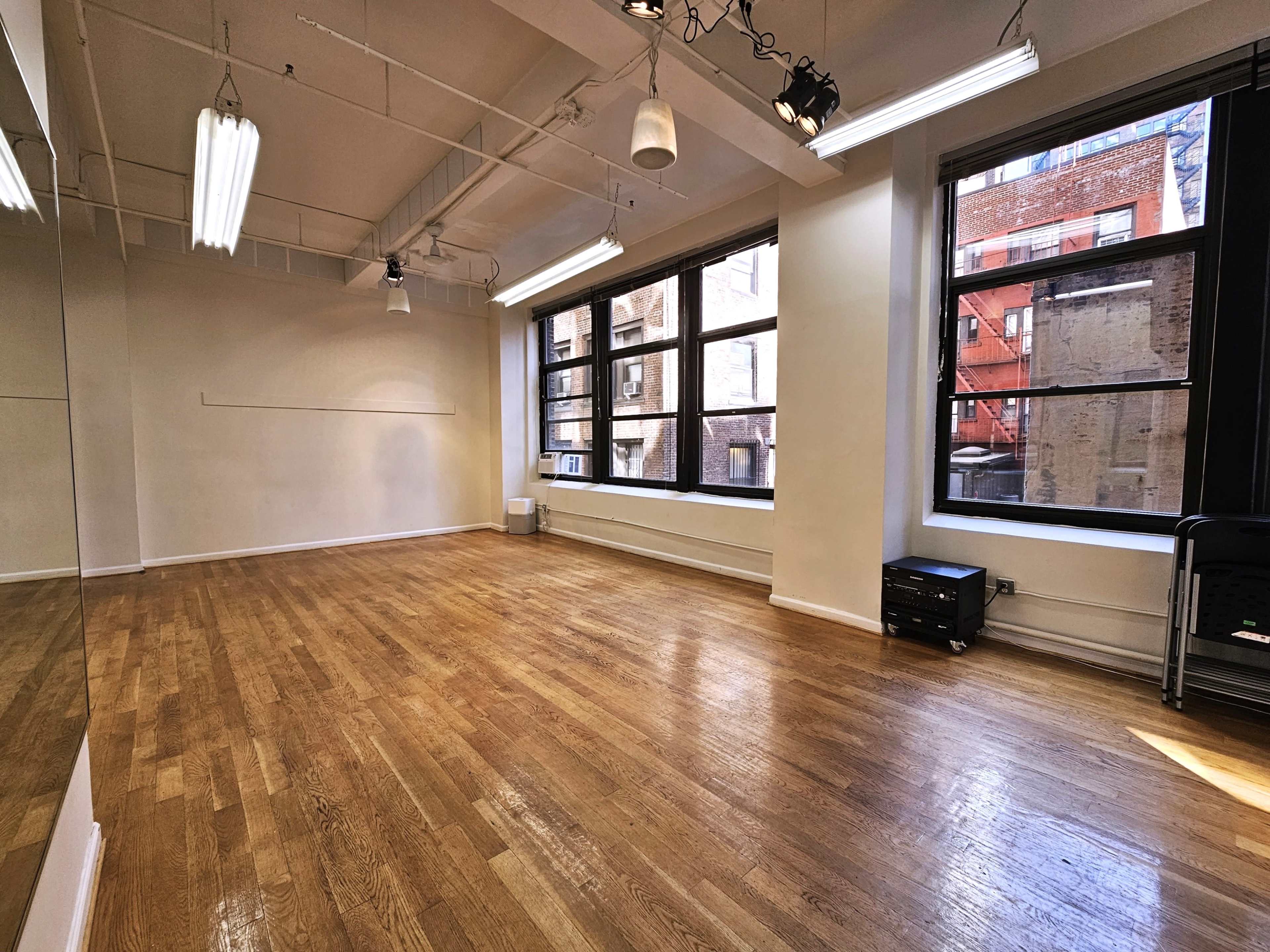 The image shows an empty studio space with hardwood floors, large windows, and bright overhead lighting.