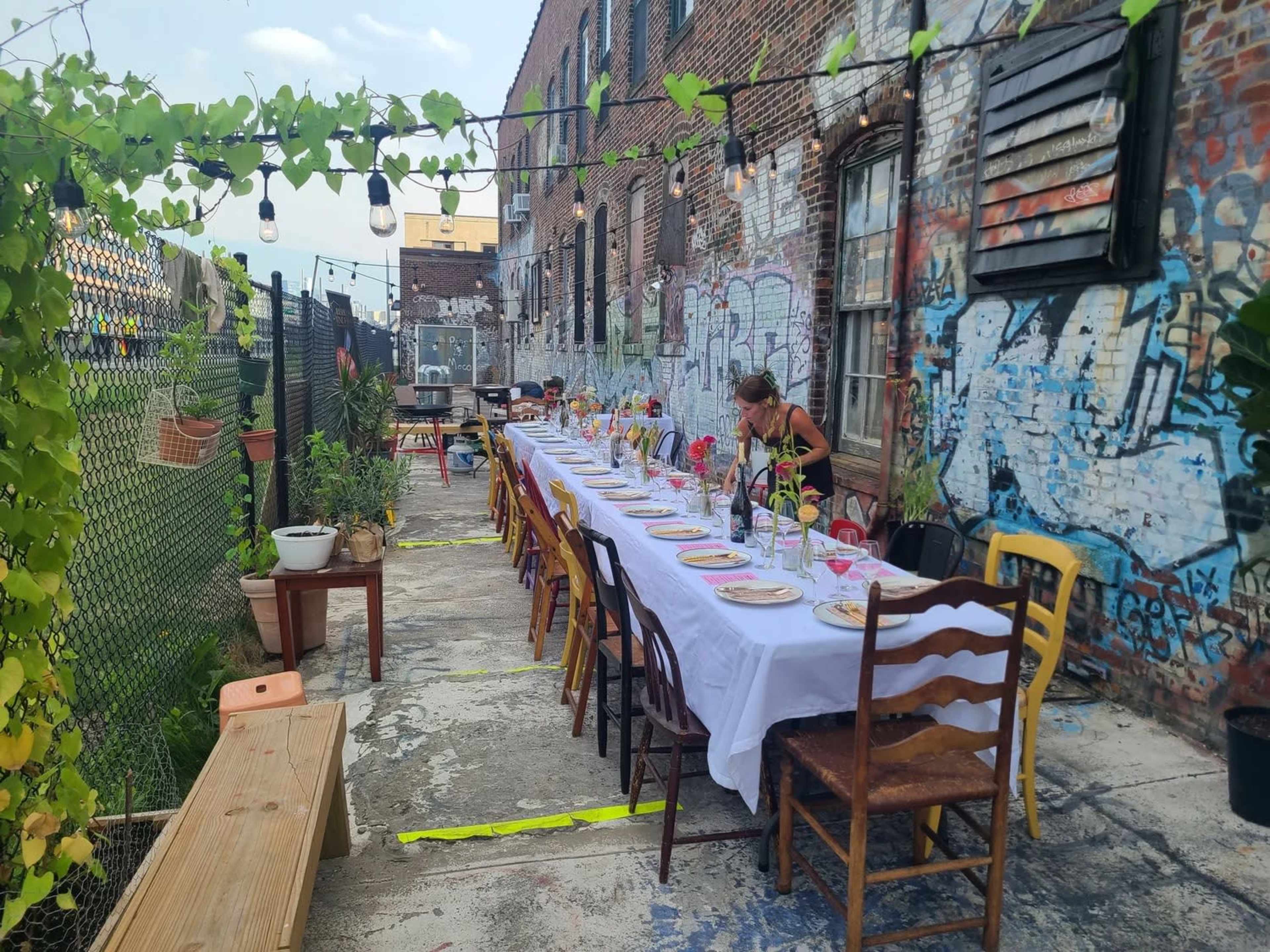 A long dining table adorned with plates and glasses is set up outside in a graffiti-covered alley, surrounded by greenery and decorative lights.
