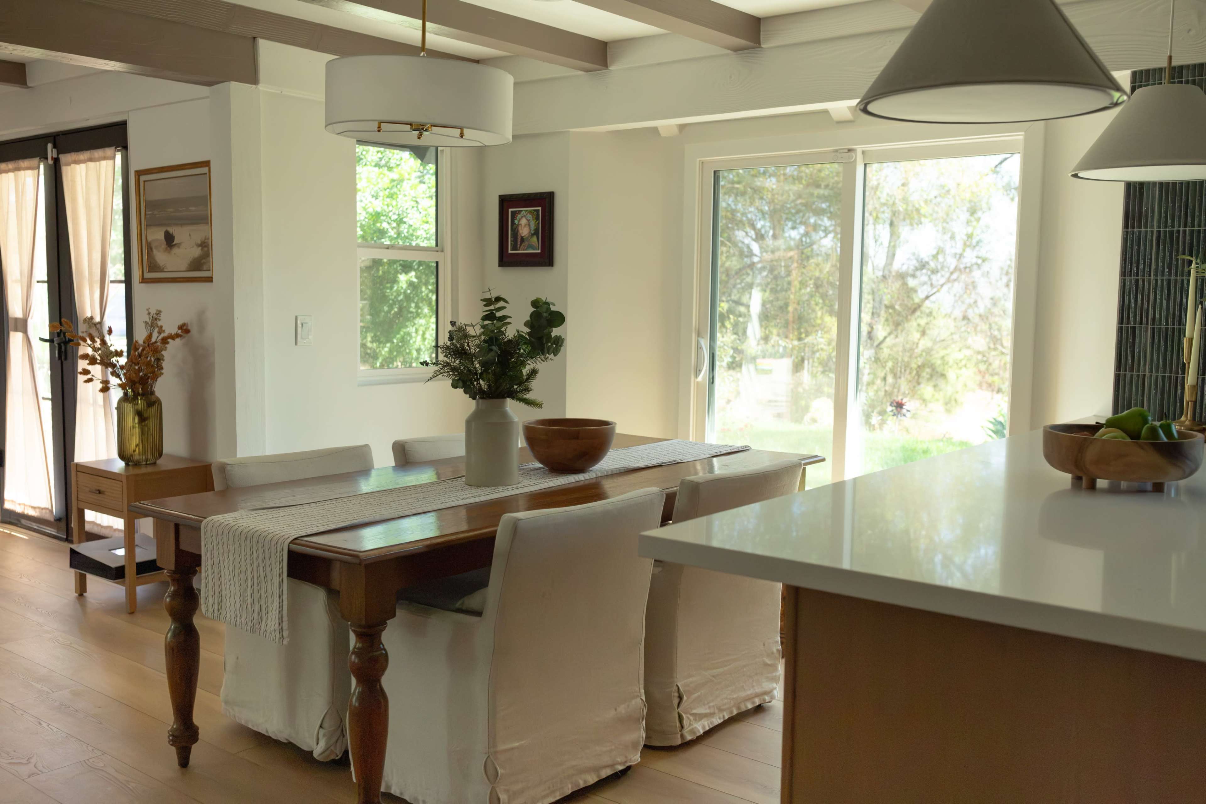 A dining area features a wooden table surrounded by white upholstered chairs, with large windows providing natural light and a view of the outdoors.