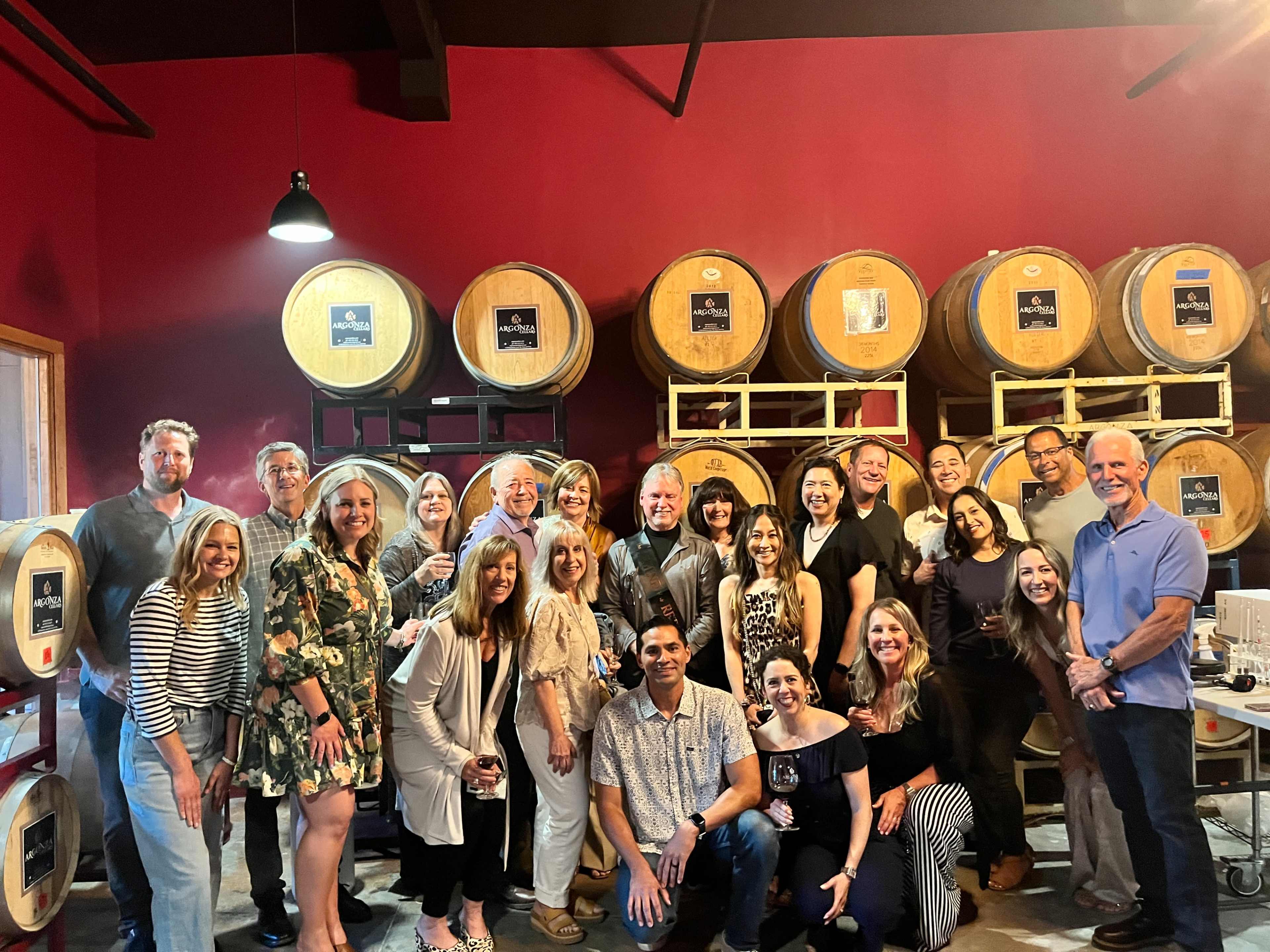 A group of around twenty people poses together in a winery or tasting room, with wooden wine barrels in the background.