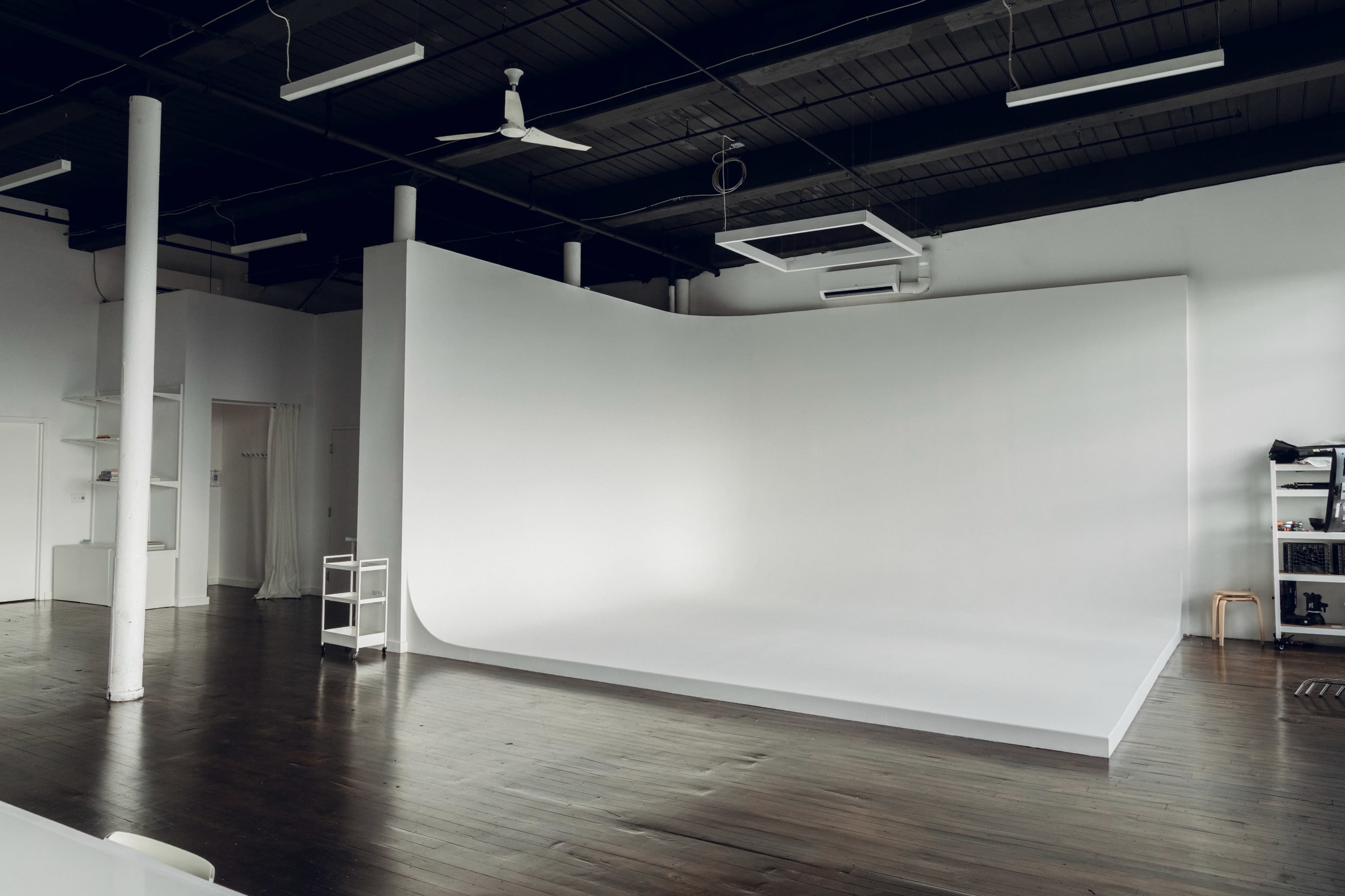 The image shows a spacious, minimalist studio with a curved white backdrop, hardwood flooring, and a few pieces of studio equipment.