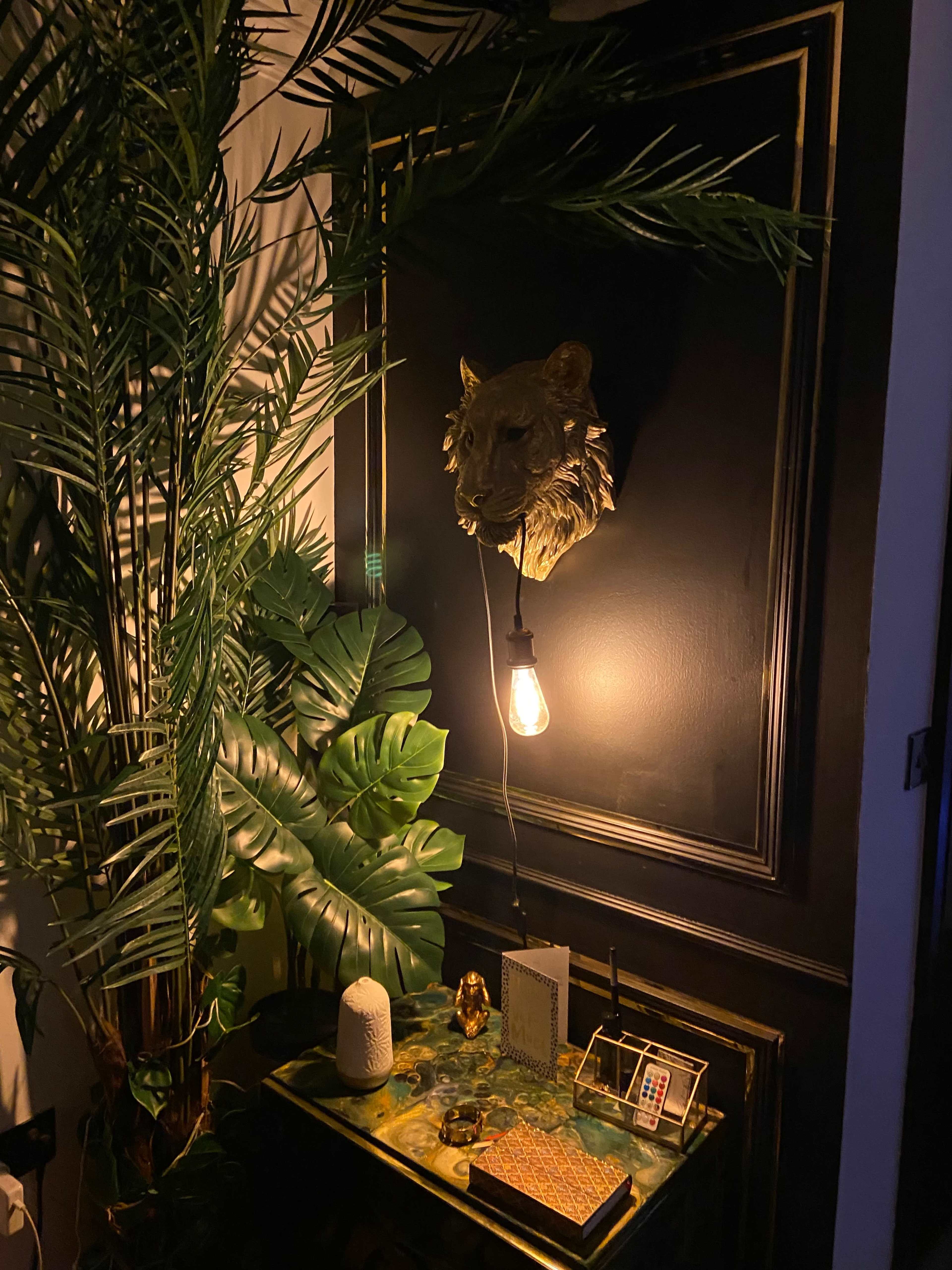 A wall decorated with a lion's head mount above a small table adorned with various items, surrounded by large green plants.