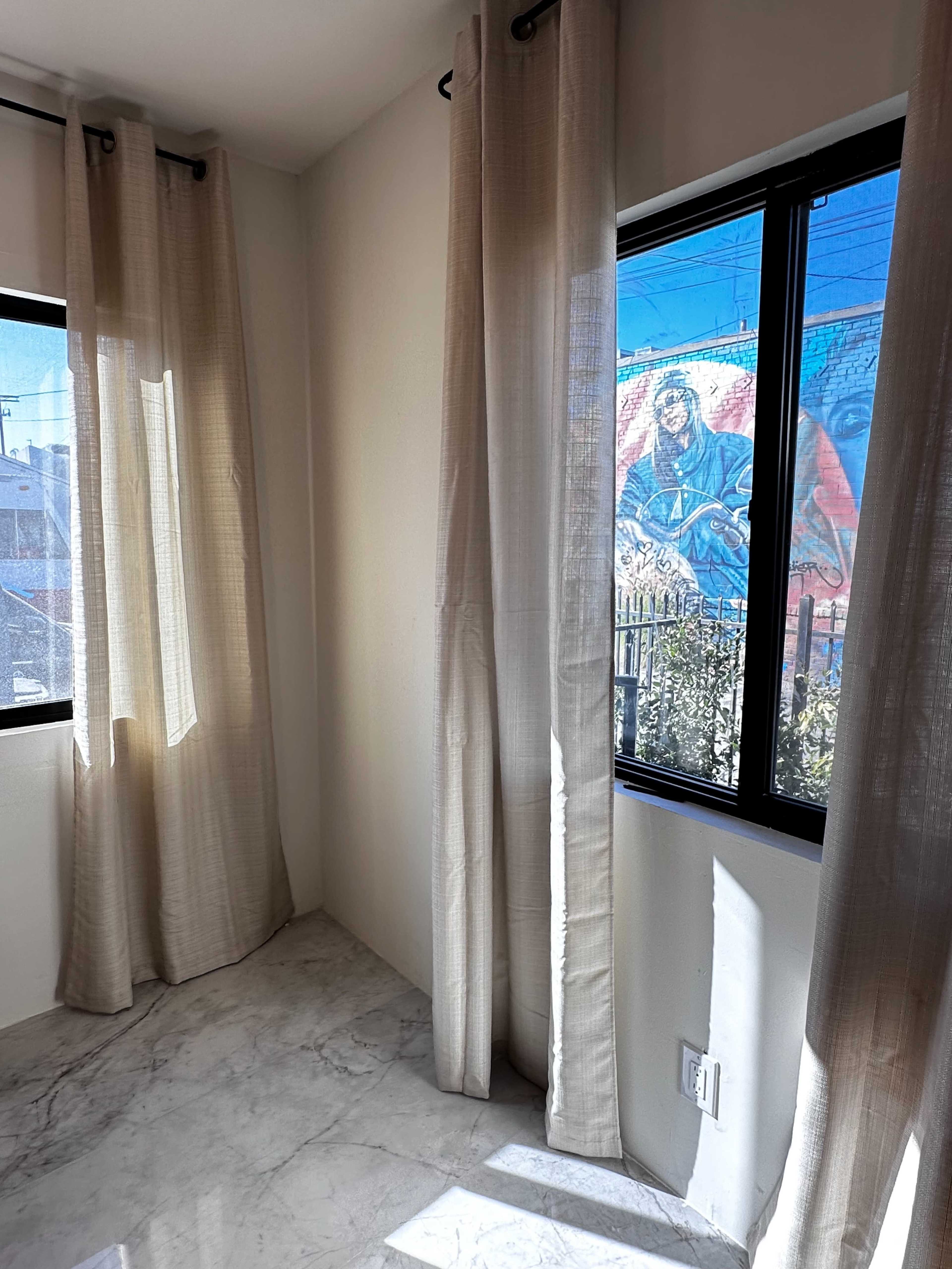 A corner of a room with large windows dressed in beige curtains, revealing a colorful mural on an exterior wall outside.