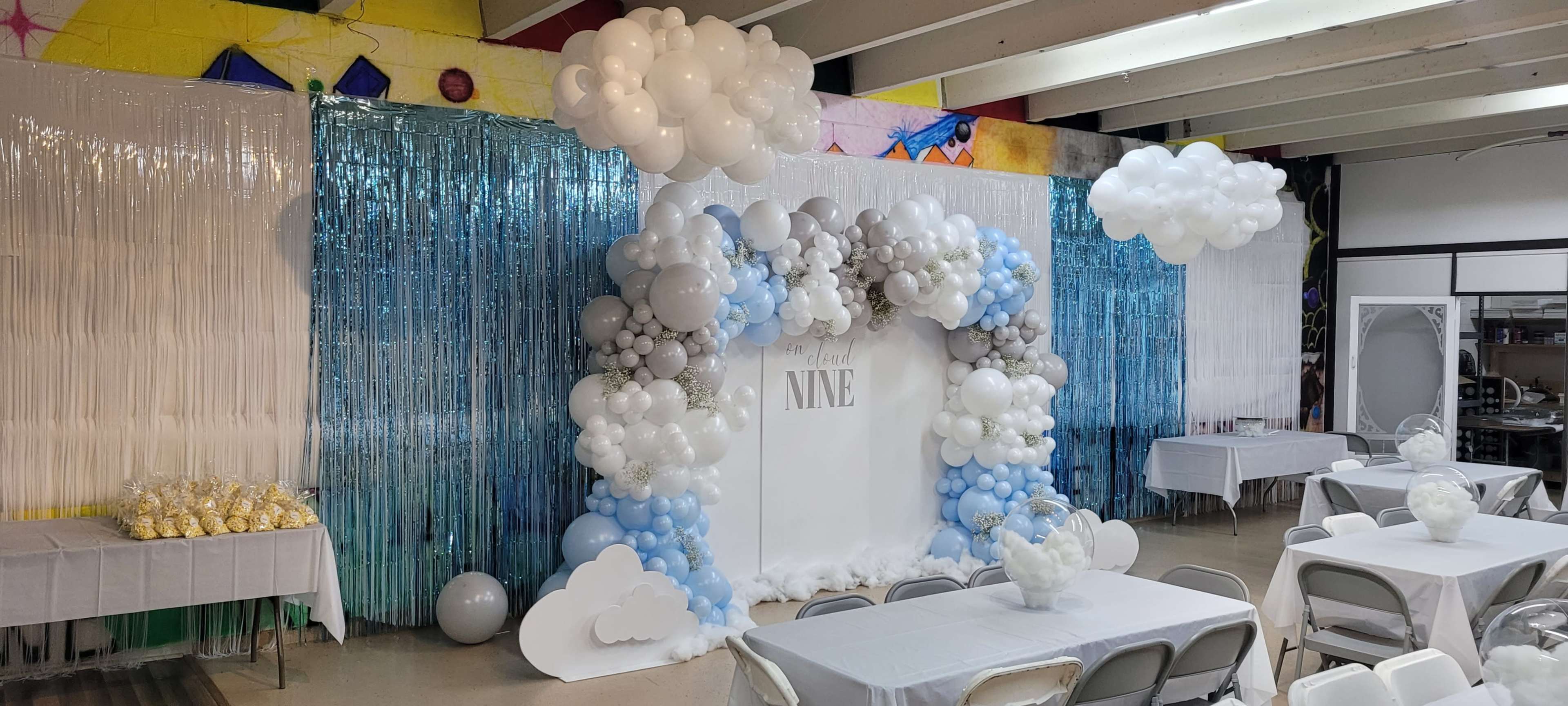 A decorated event space featuring a balloon arch in blue, white, and gray, with a backdrop that reads "Happy NINE," surrounded by tables covered with white tablecloths.