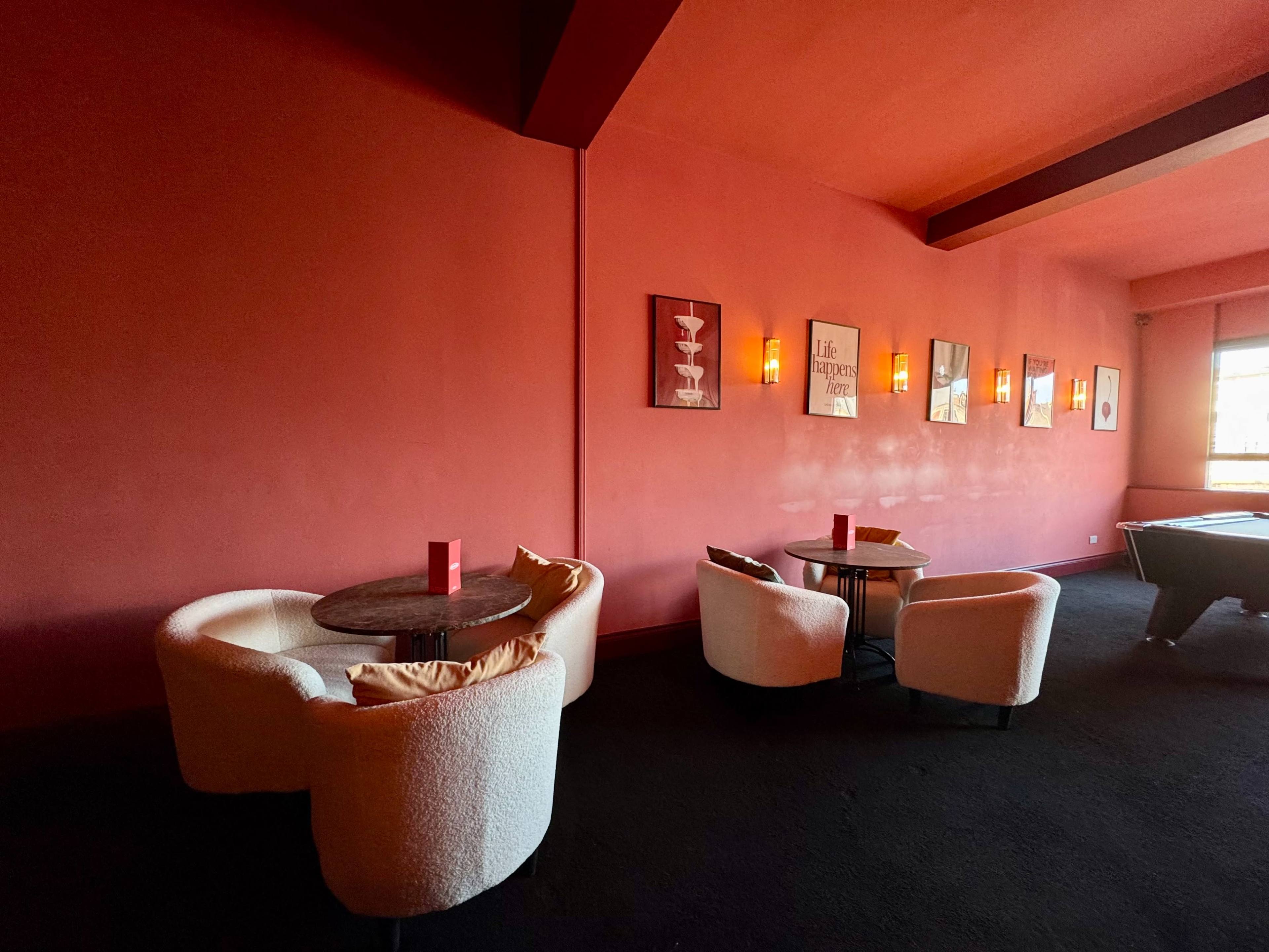 A dimly lit room features several circular chairs and small tables against a red wall, with framed artwork and soft lighting.