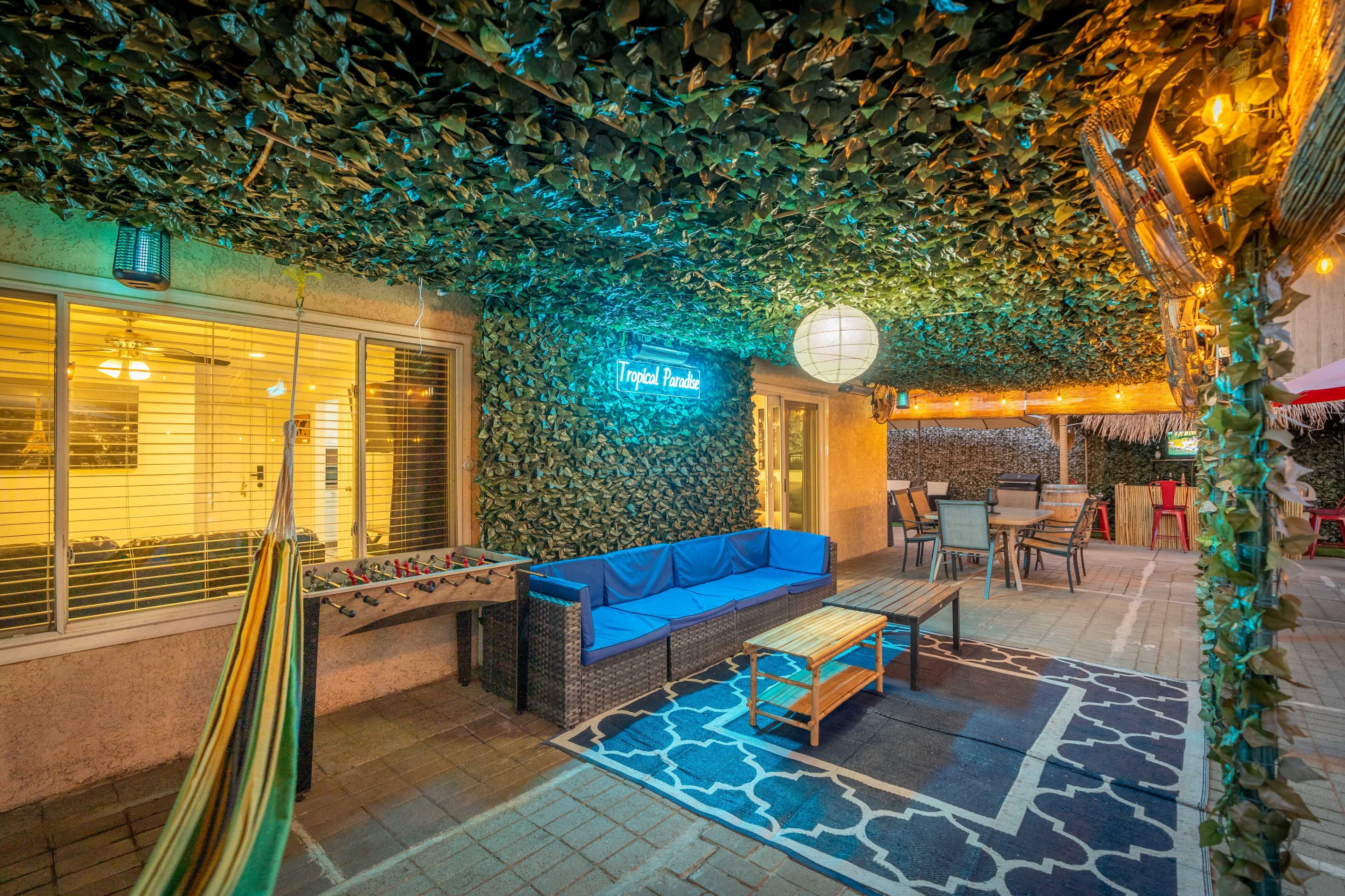 A patio area with a blue couch, a wooden coffee table, and a hammock, all surrounded by greenery overhead and string lights.