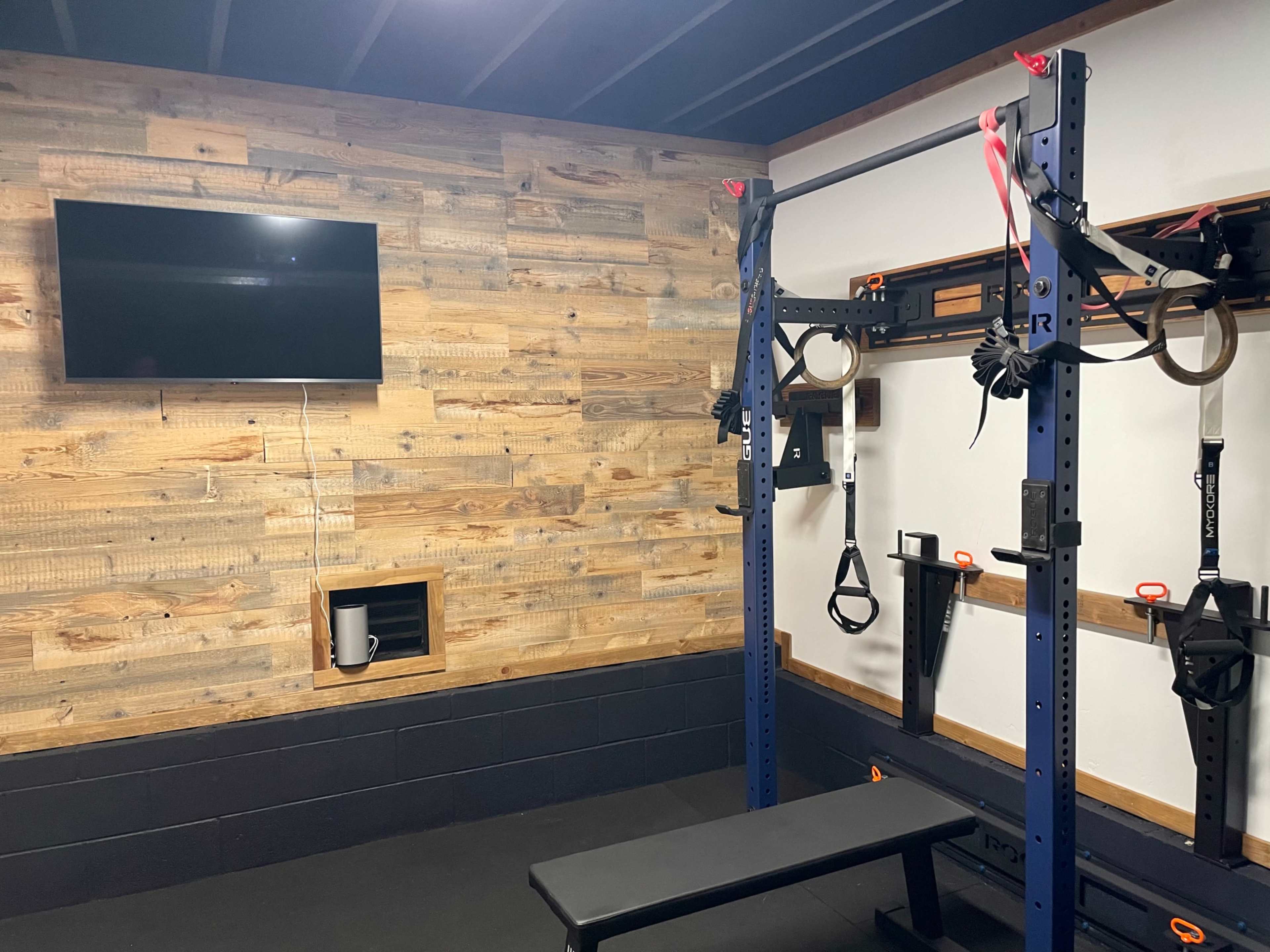 A home gym with a weight bench, a squat rack mounted on a wooden wall, and a television above.
