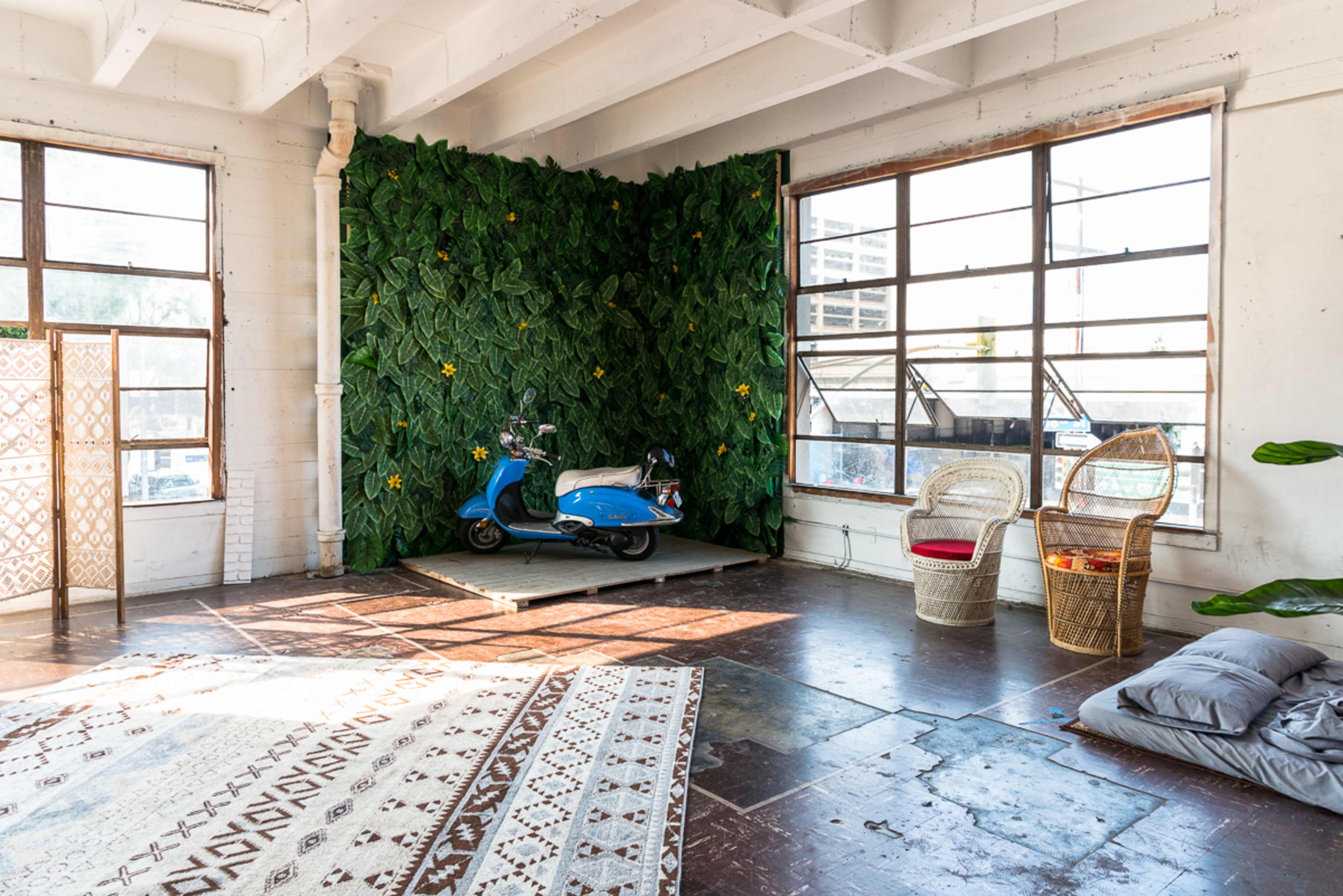 A spacious room with large windows, featuring a green wall of foliage, a blue scooter, wicker chairs, and a patterned rug.