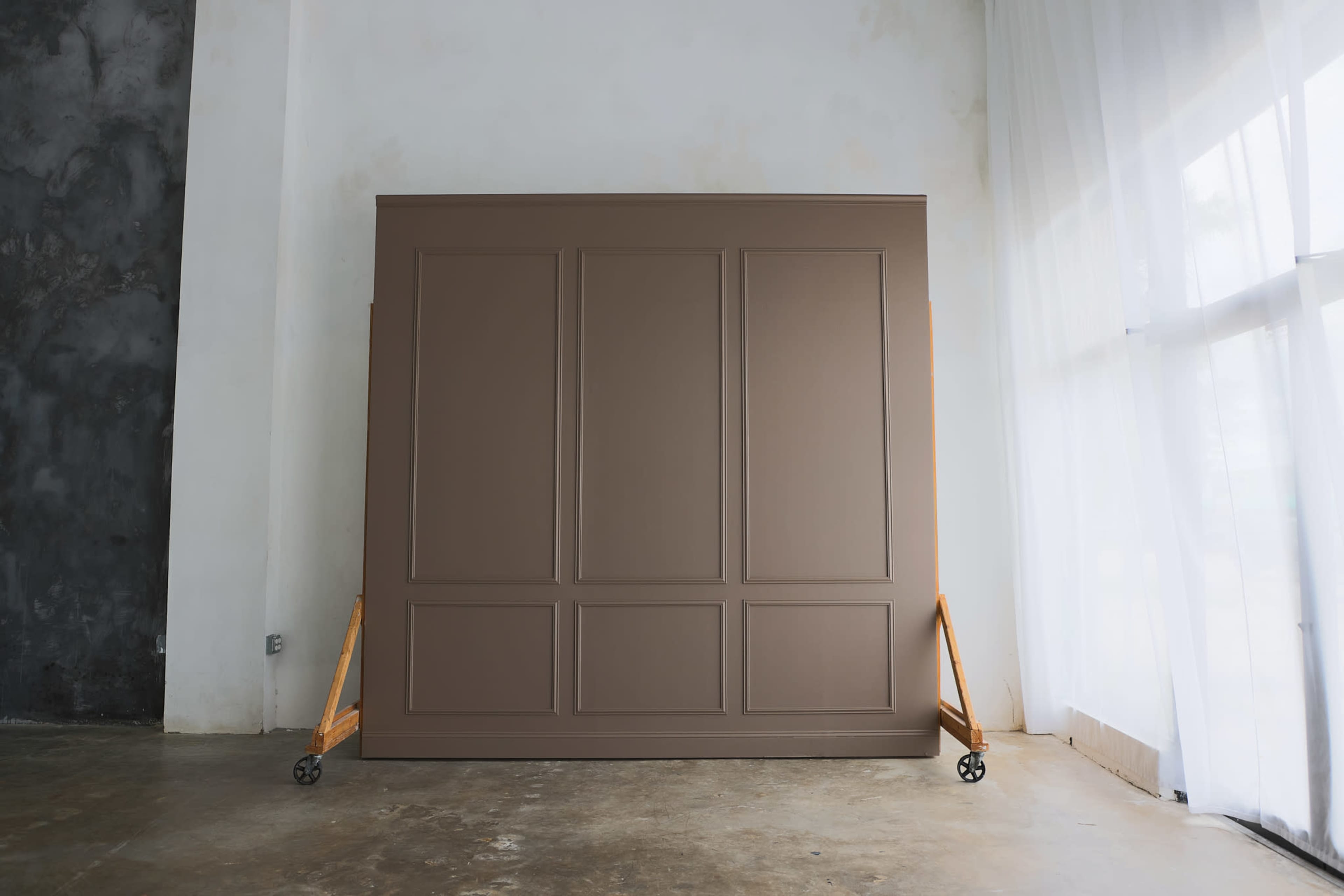 A large brown panel with a geometric design is positioned upright on wheels in a minimalist interior with white drapes and a concrete floor.