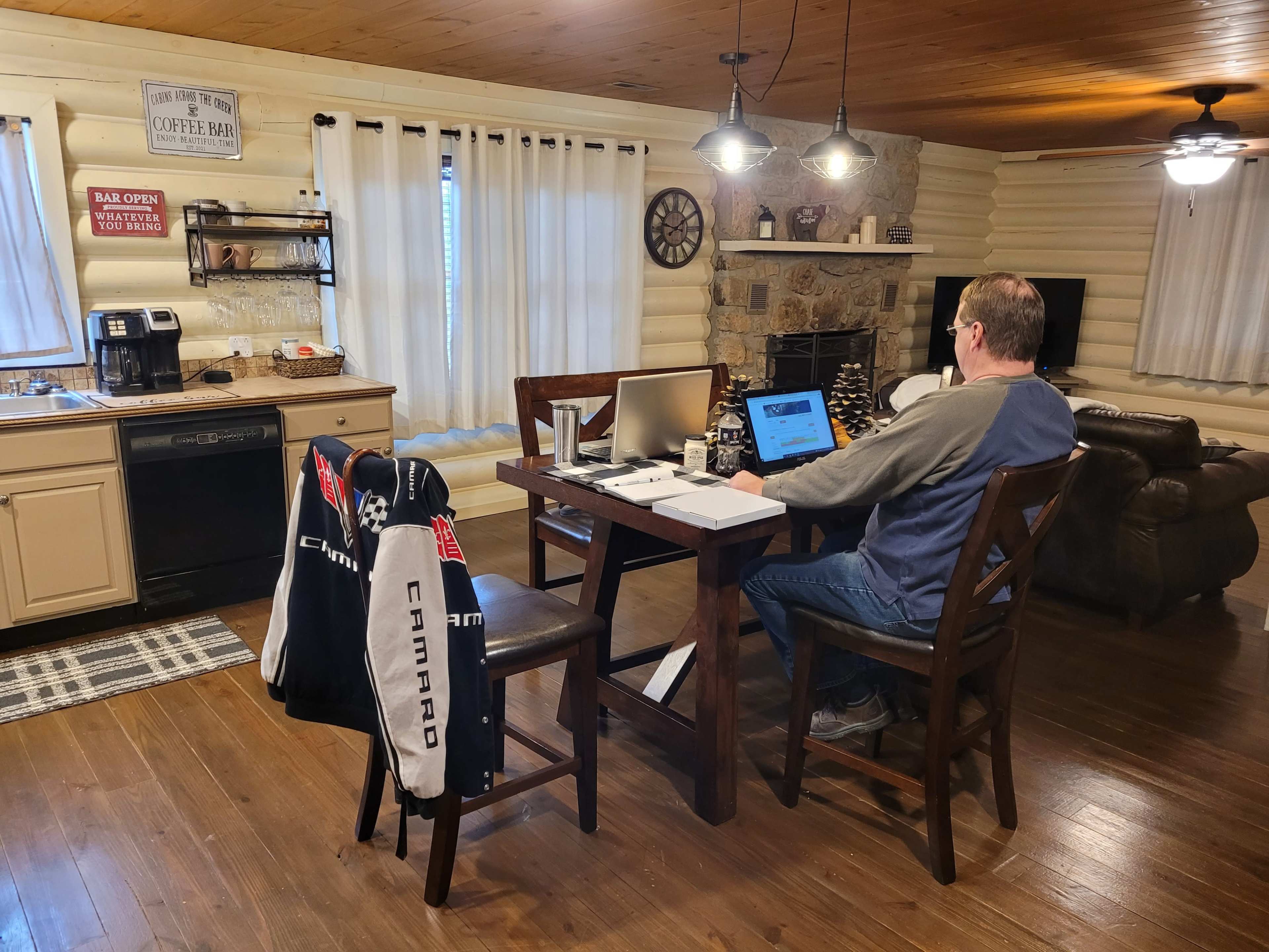 A person is working at a wooden table in a cozy kitchen with log cabin walls, a coffee maker, and a stone fireplace.