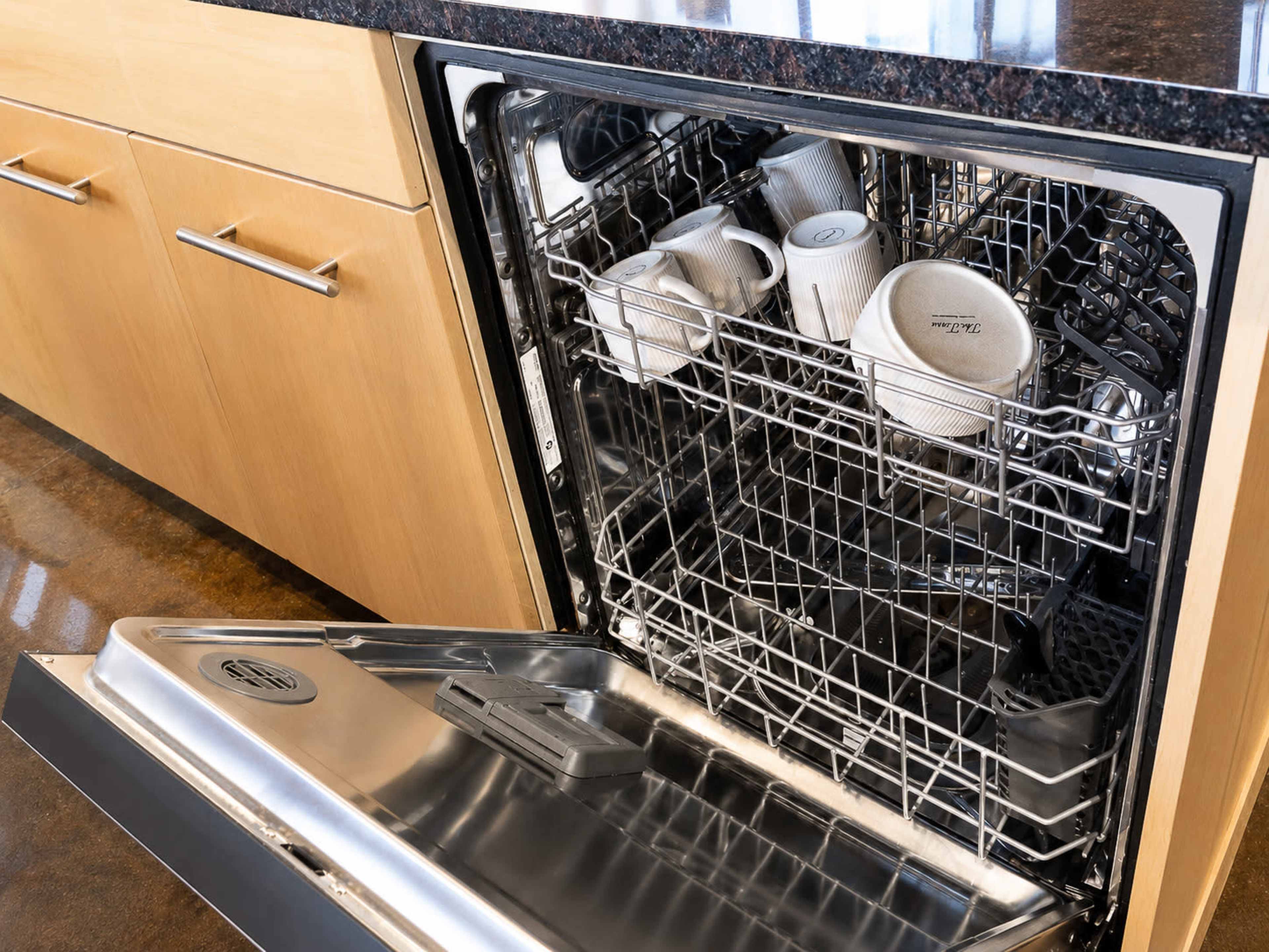 The image shows an open dishwasher filled with several white cups placed on the top rack, set against a backdrop of wooden kitchen cabinetry.