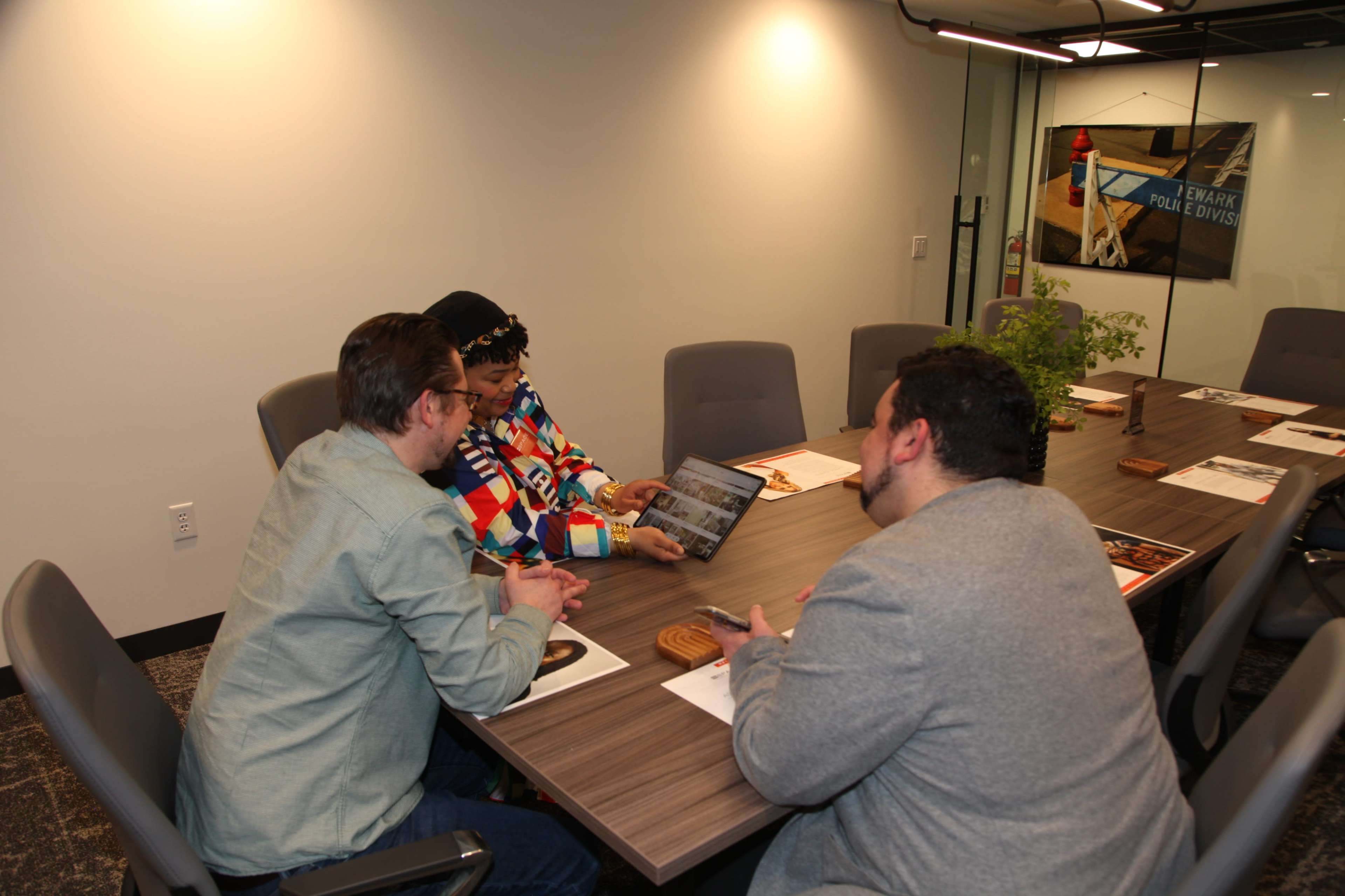 Three individuals are seated at a conference table, reviewing materials on a tablet and discussing in a modern meeting room.
