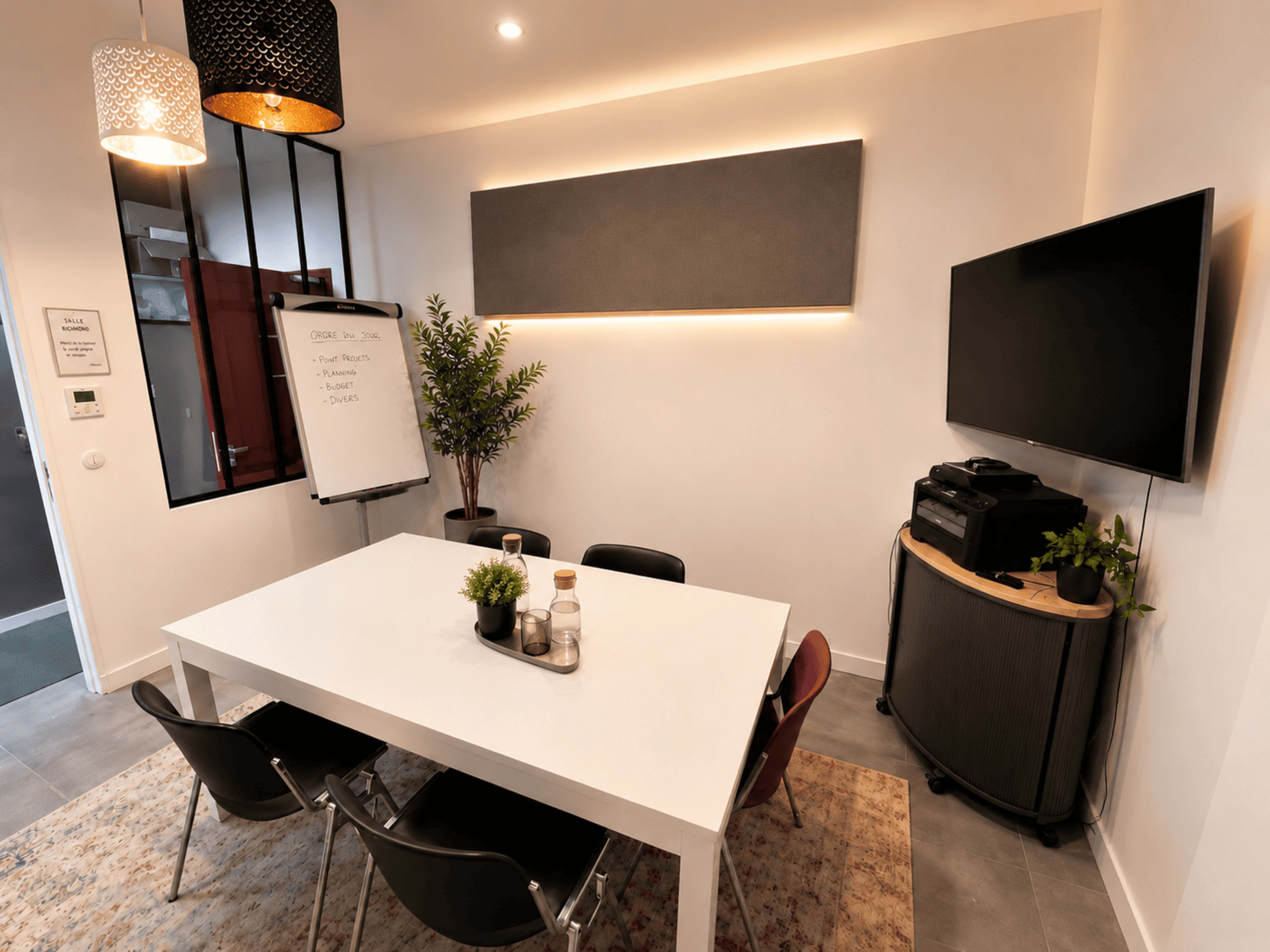 A modern meeting room features a white table surrounded by black chairs, a wall-mounted TV, a plant, and a whiteboard illuminated by pendant lights.