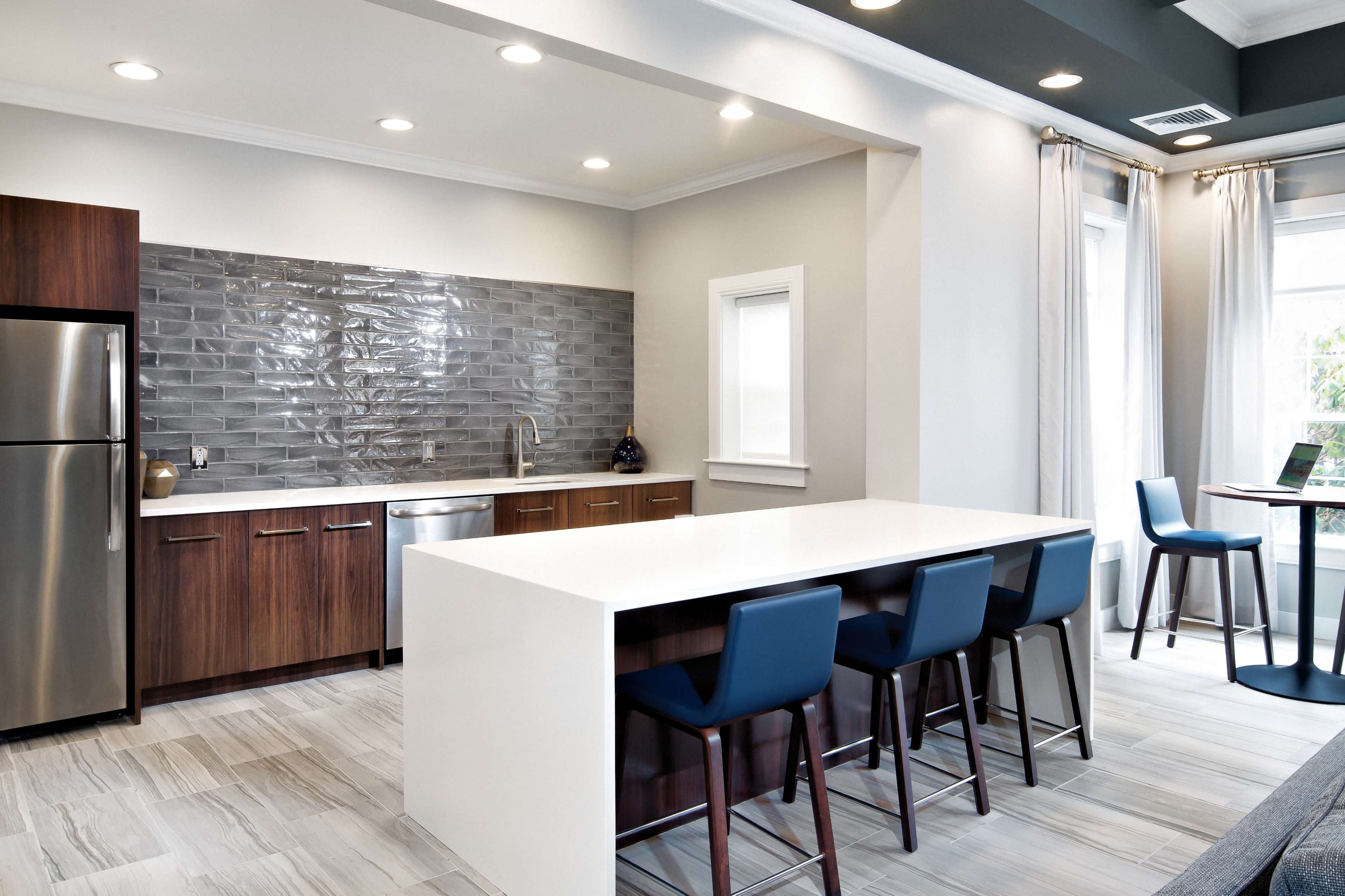 A modern kitchen features gray tile backsplash, dark wood cabinets, a stainless steel refrigerator, and a large white island with blue bar stools.