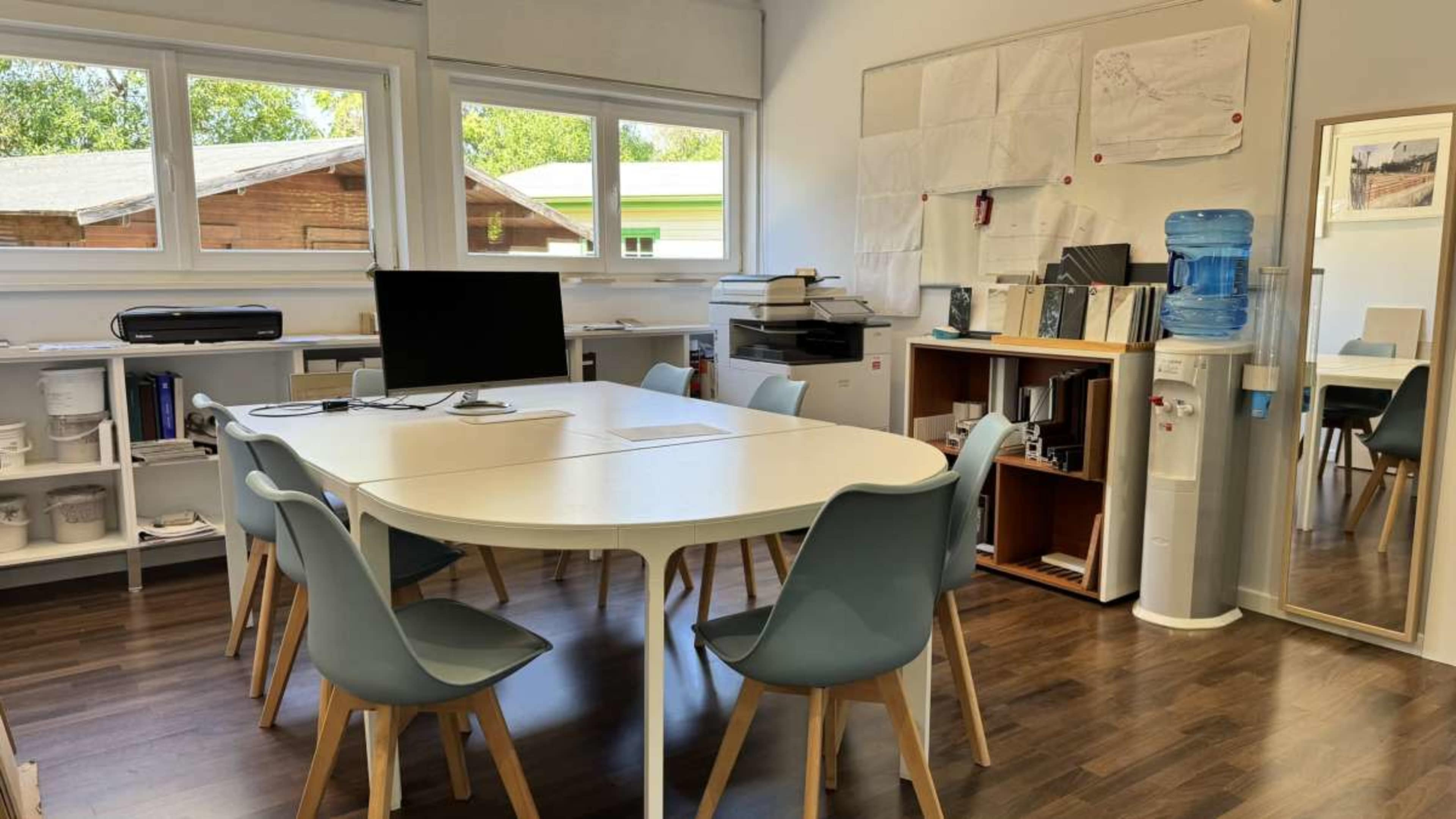 The image shows an office space with a round table surrounded by six blue chairs, a computer monitor, a printer, and shelves filled with documents and supplies.