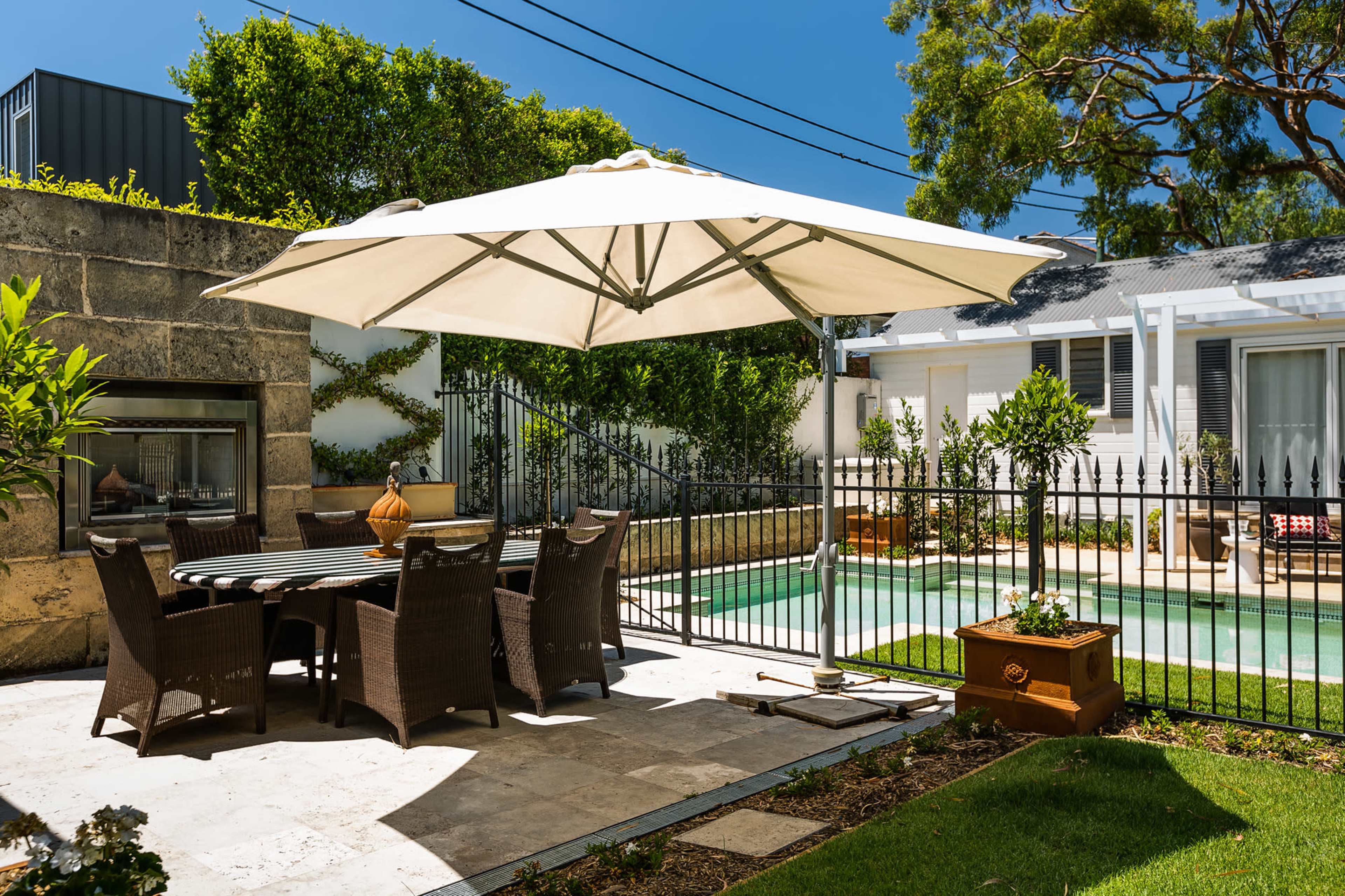 A patio area features a large umbrella over a dining table surrounded by chairs, with a fenced pool and a house in the background.