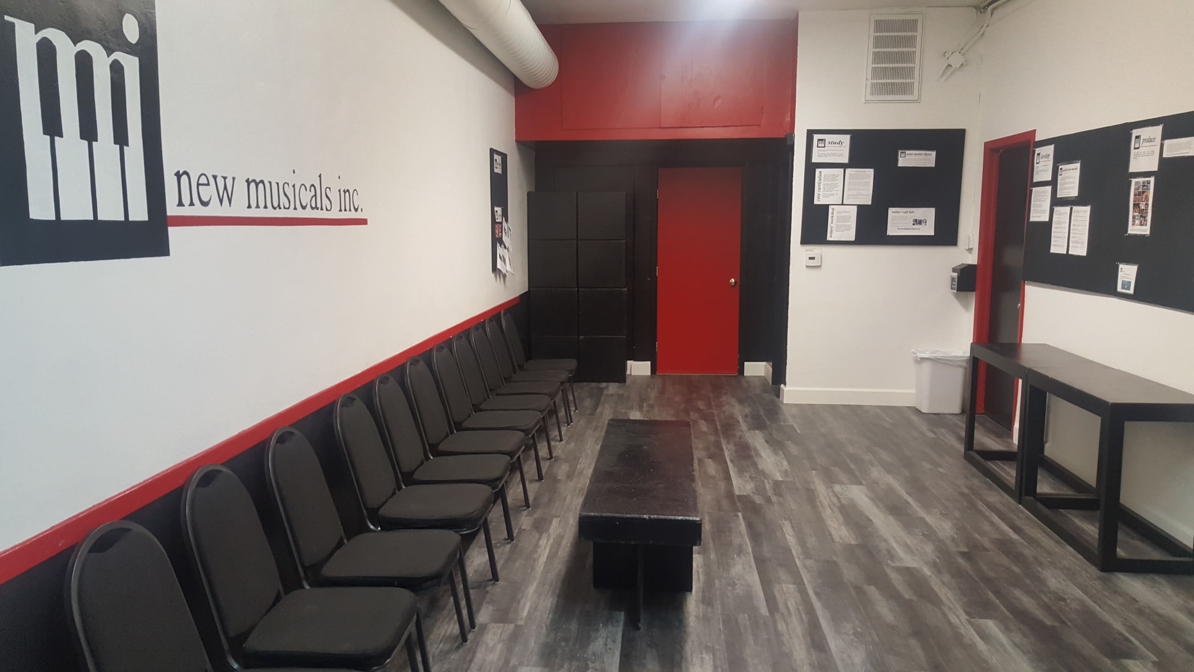The image shows a waiting area for New Musicals Inc., featuring a row of chairs along the wall opposite a long table and a red door.