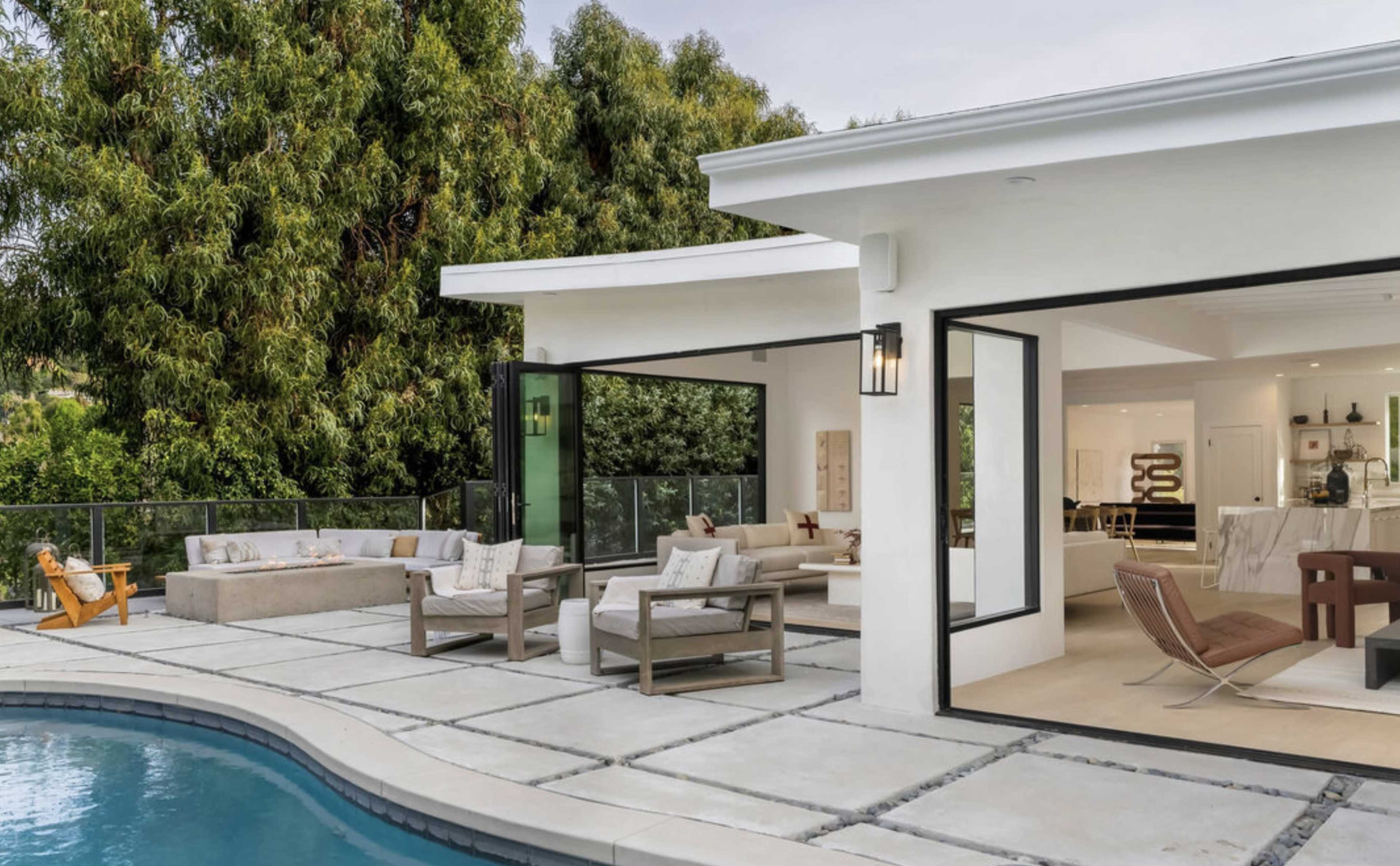 A modern home features an open patio area with outdoor seating beside a swimming pool, surrounded by greenery.