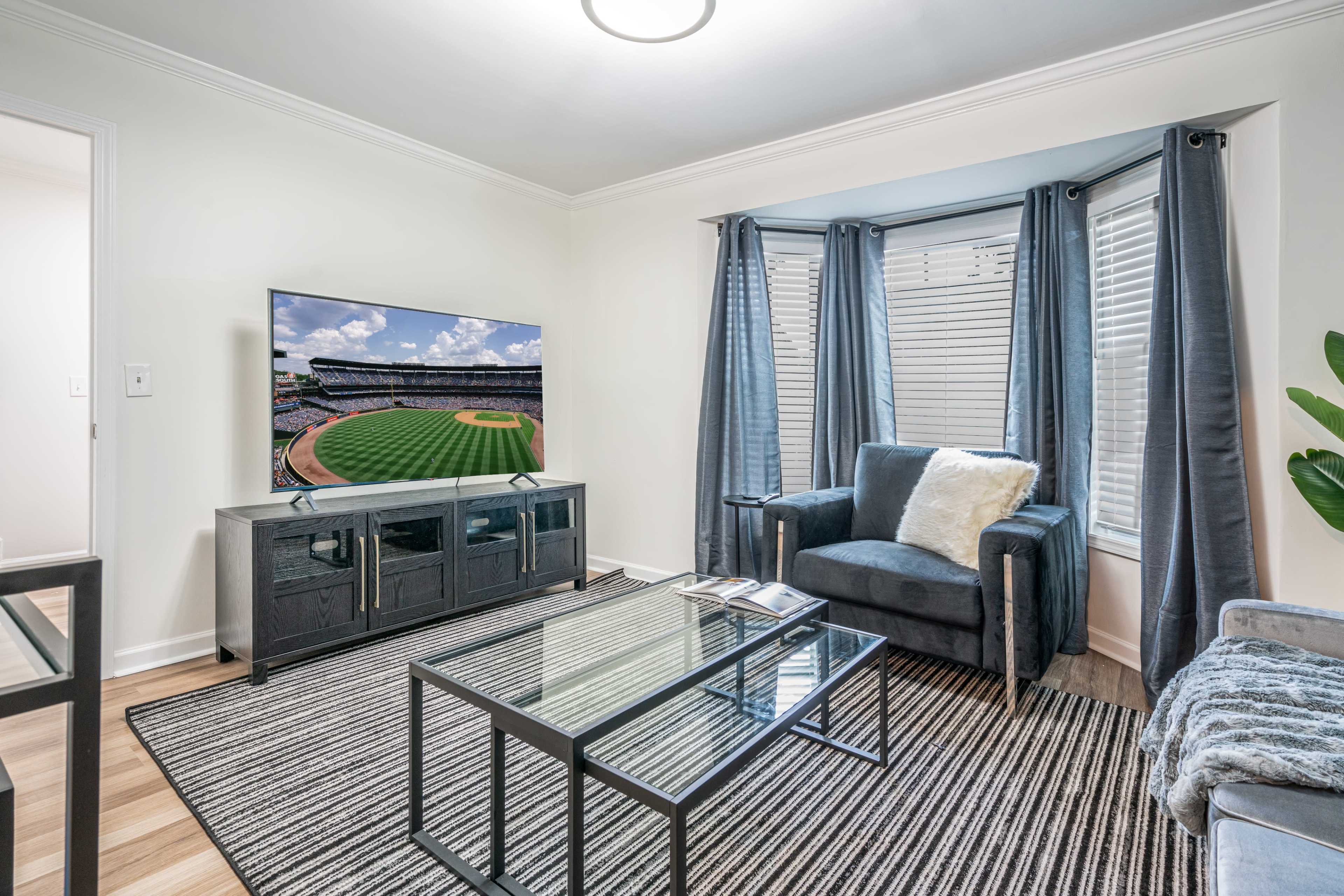 A living room features a television displaying a baseball field, flanked by large windows with curtains, a glass coffee table, and two armchairs.