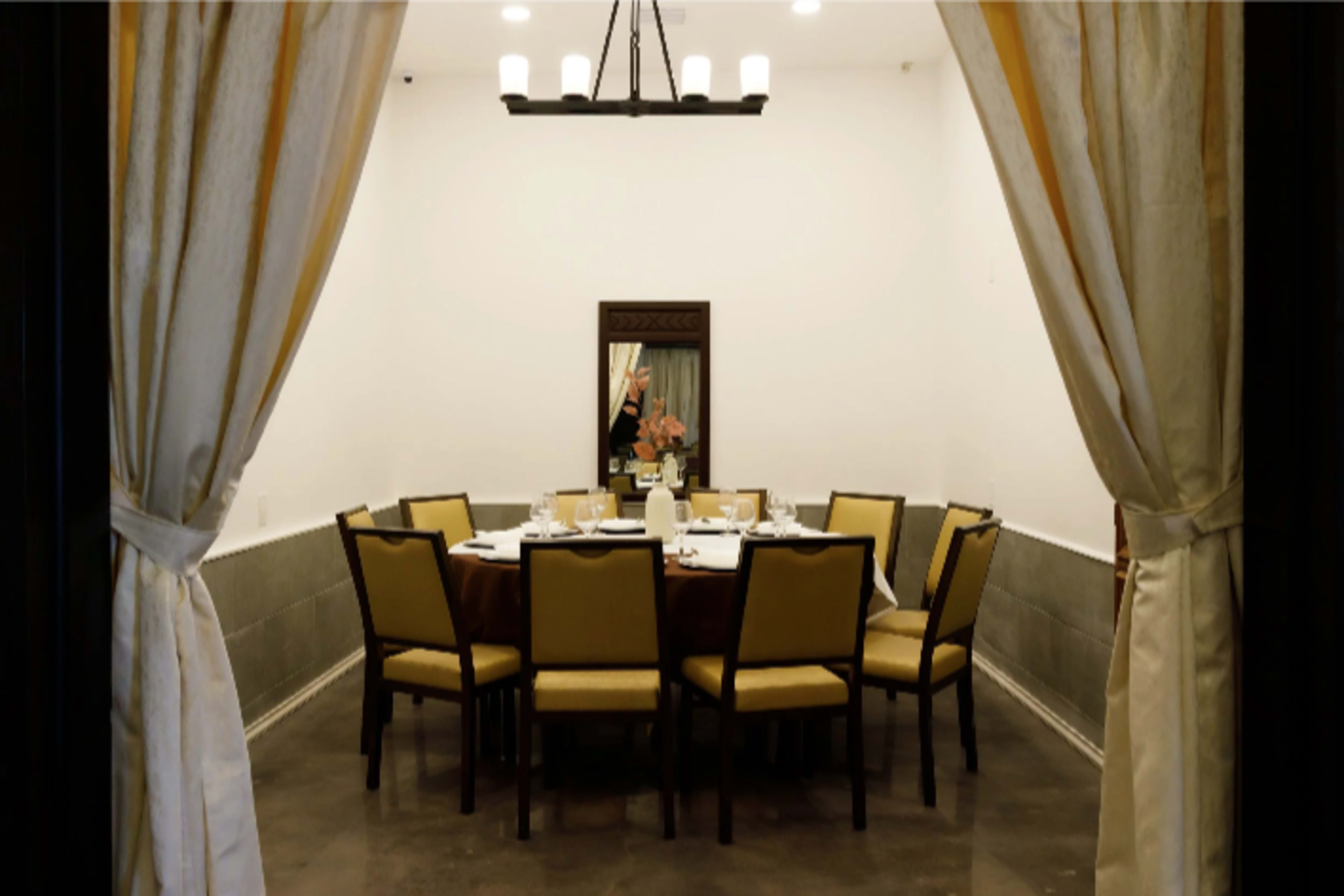 A private dining room features a large round table set with diningware and surrounded by chairs, framed by white curtains.
