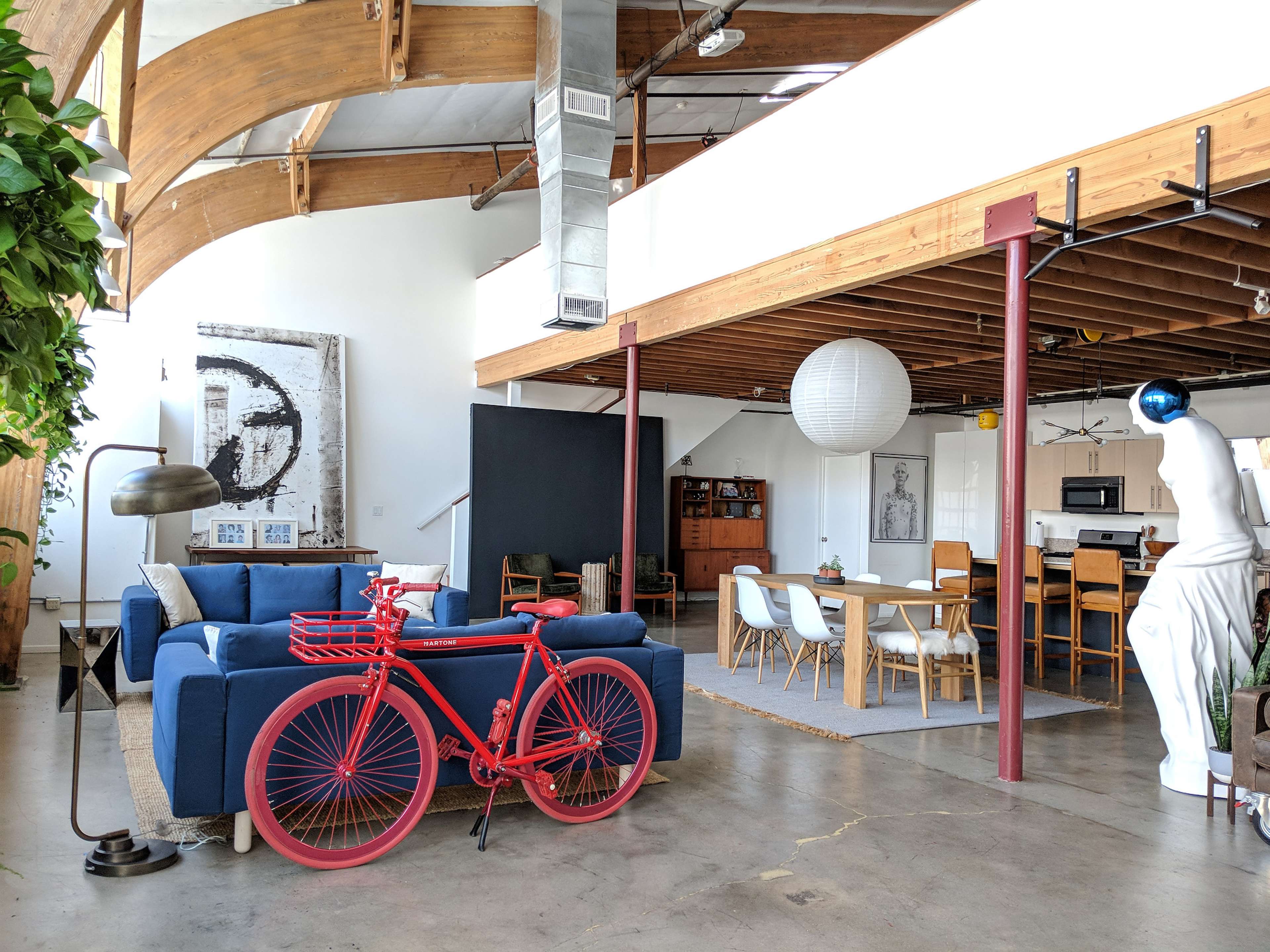 A spacious loft-style living area with a blue sofa, a red bicycle, and a dining table surrounded by chairs, all under wooden beams and bright lighting.