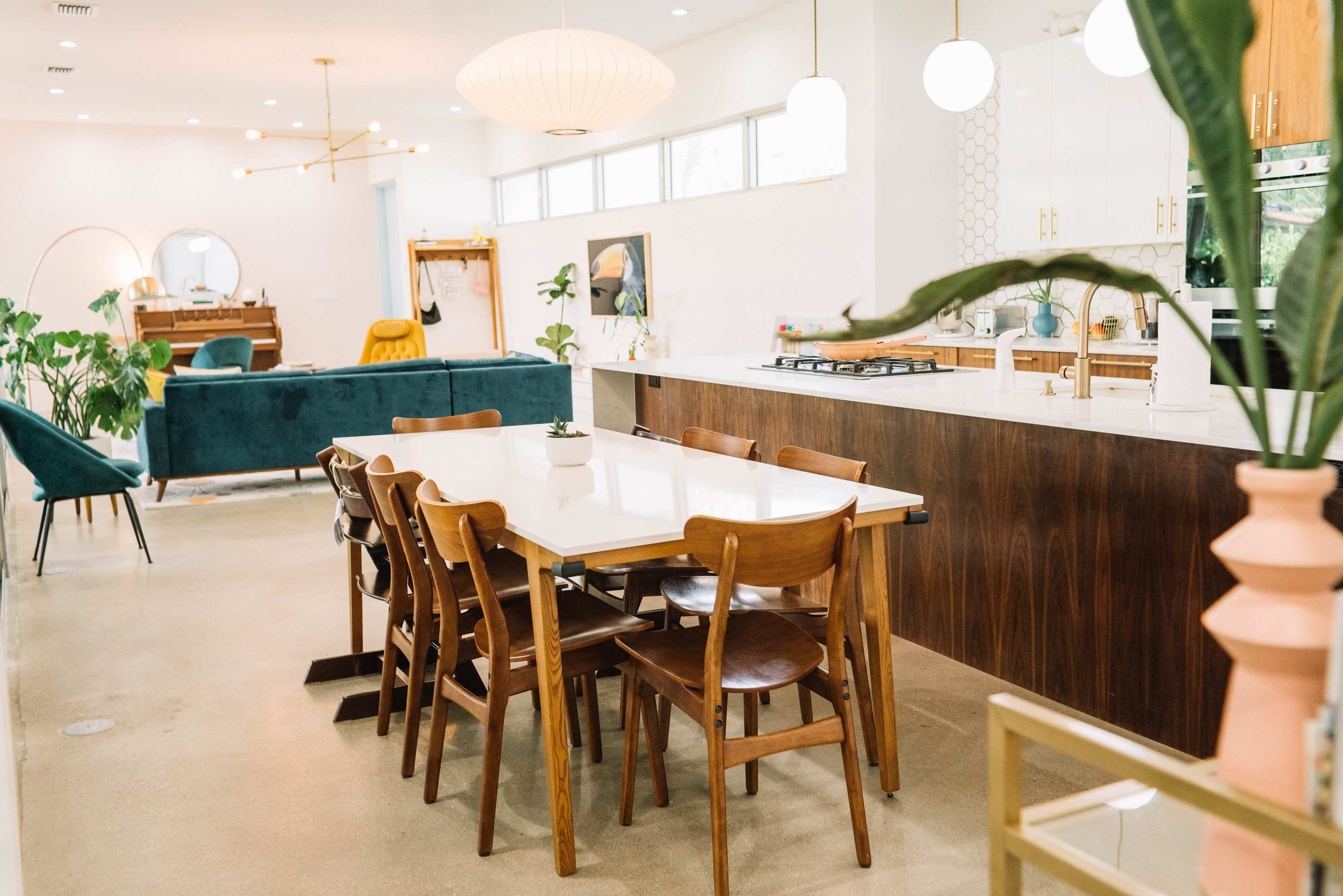 The image shows a spacious modern kitchen and dining area with a large table surrounded by wooden chairs, featuring an open layout and large windows.