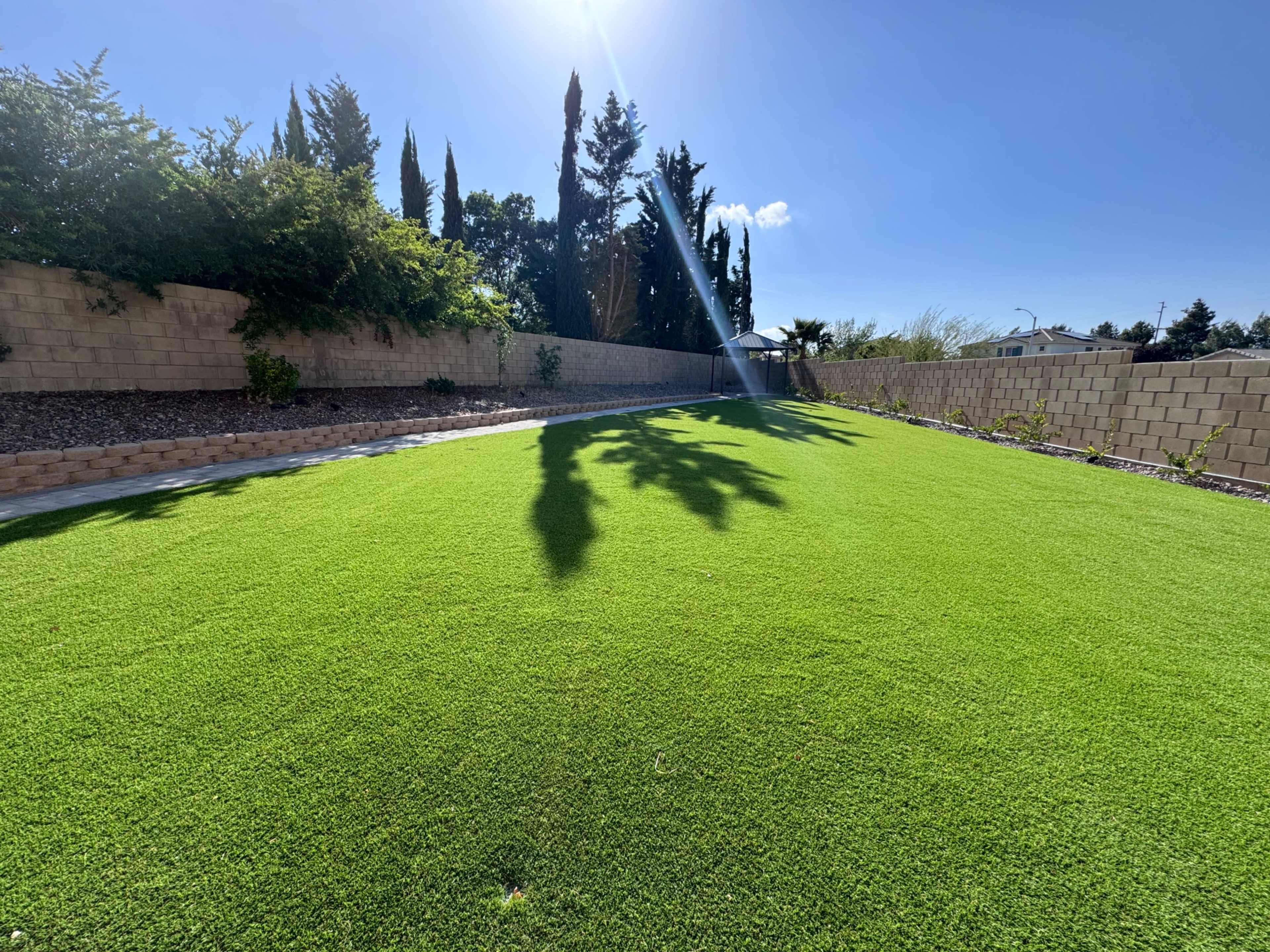 A green artificial lawn is surrounded by a low stone wall and various trees under a clear blue sky.