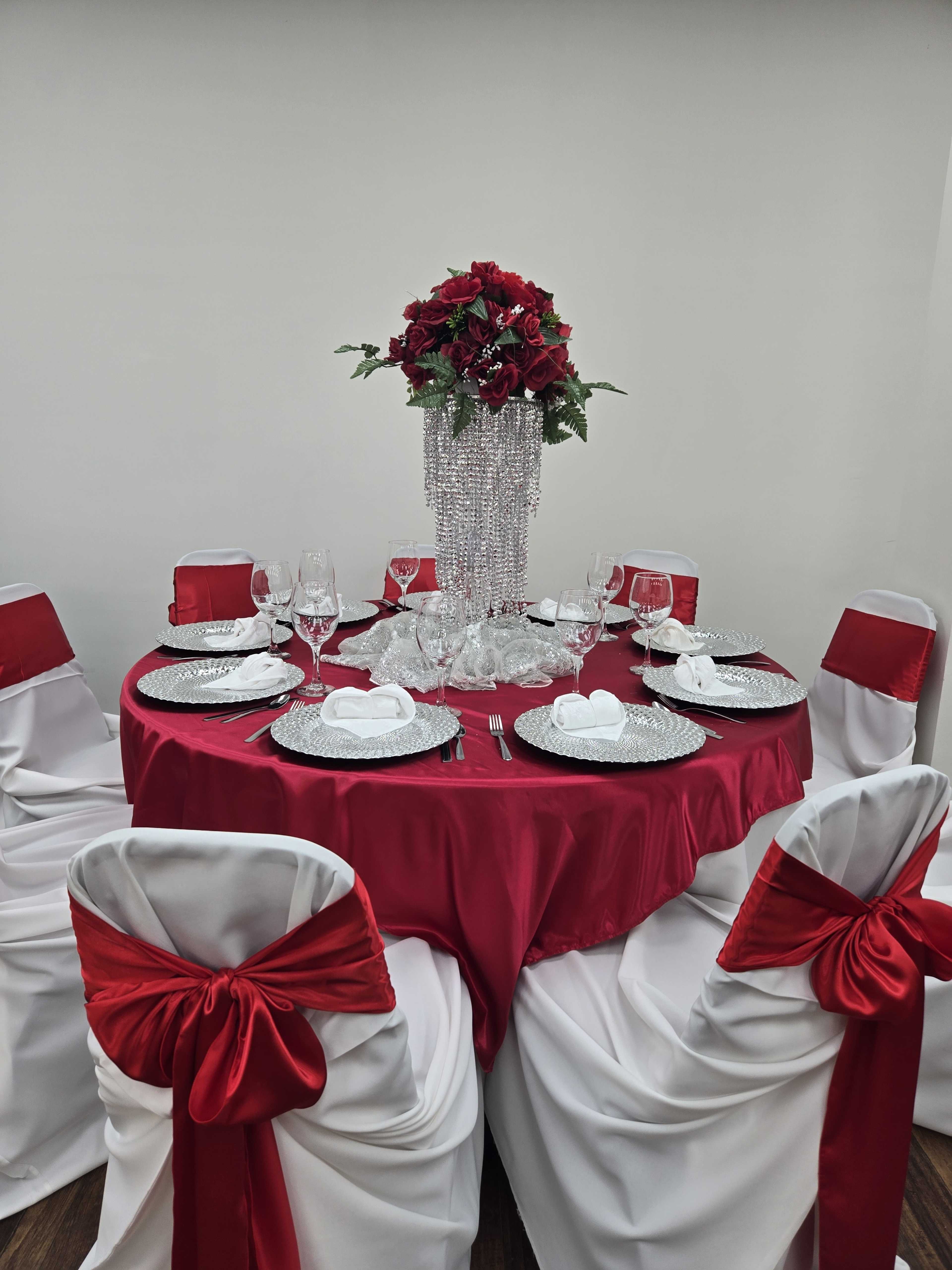 A round table is set for a formal dining arrangement, featuring a red tablecloth, crystal glassware, and a tall floral centerpiece adorned with red roses.