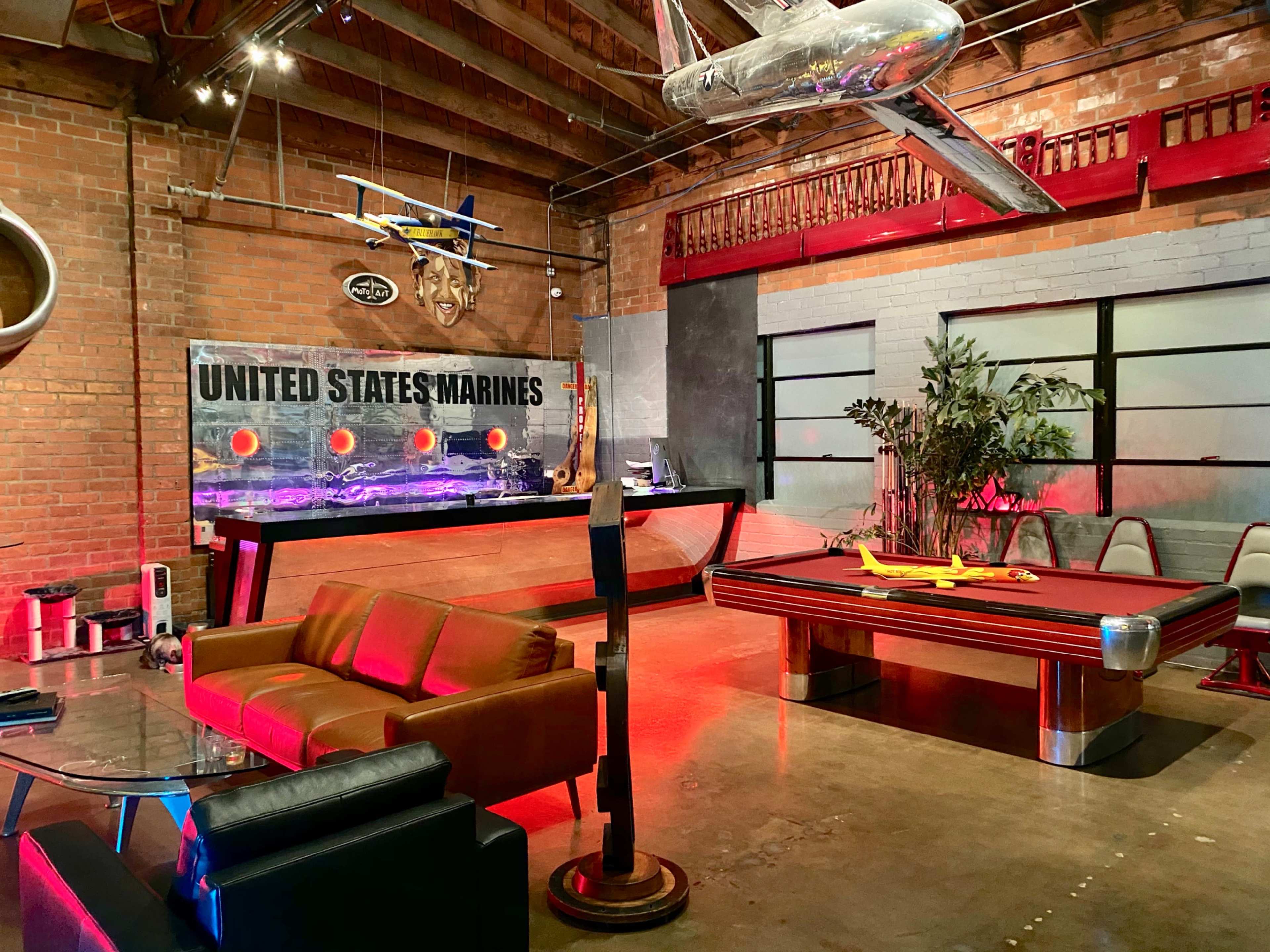 The image shows a modern lounge area with a brown sofa, a pool table, and military-themed decor, including a wall displaying "UNITED STATES MARINES."
