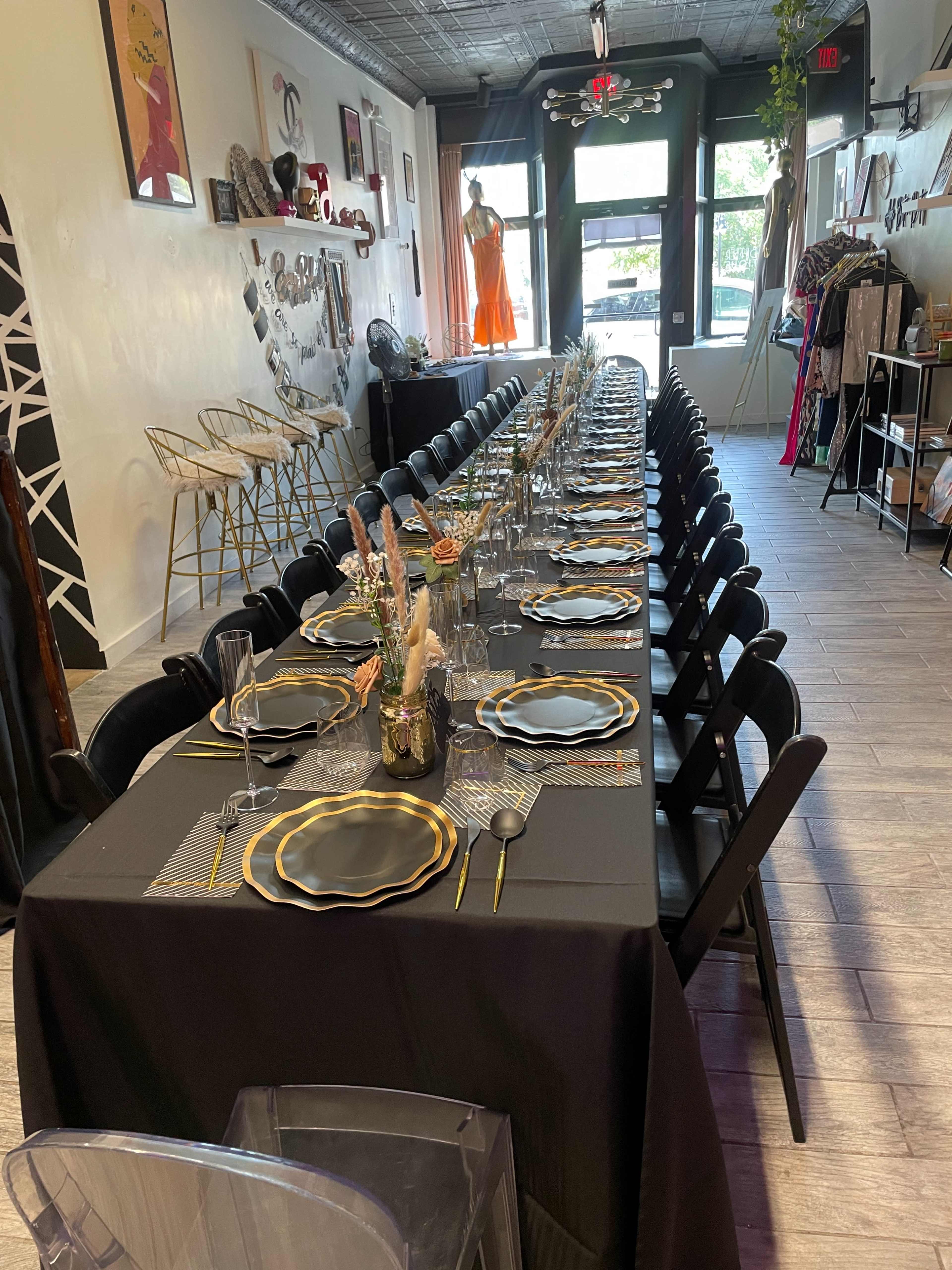 A long, elegantly set table with black tablecloth and gold-accented dinnerware occupies a stylishly decorated dining space.